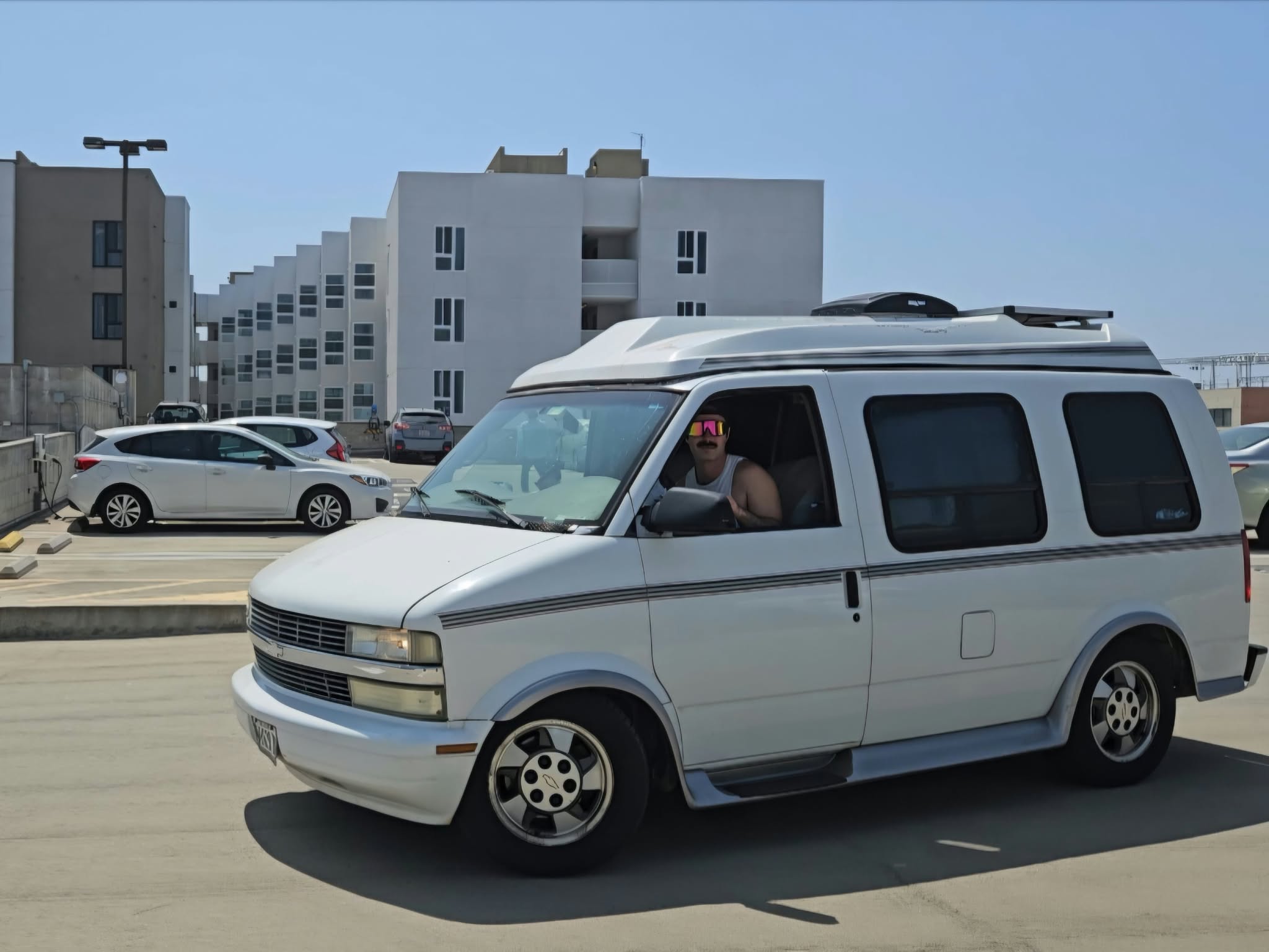 White van driving on street with buildings and parked cars in background.