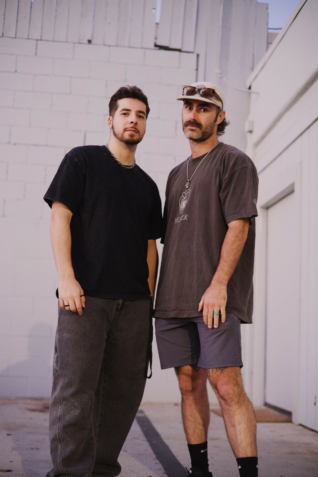 Two men stand outdoors against a white wall, one in a black t-shirt and the other in a gray t-shirt and shorts.