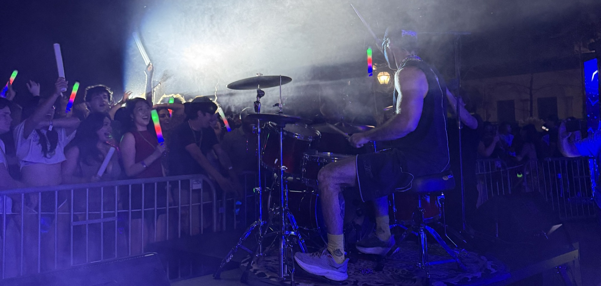 Musician playing drums on stage with audience holding glow sticks, dim lighting, and fog effects, lively atmosphere.