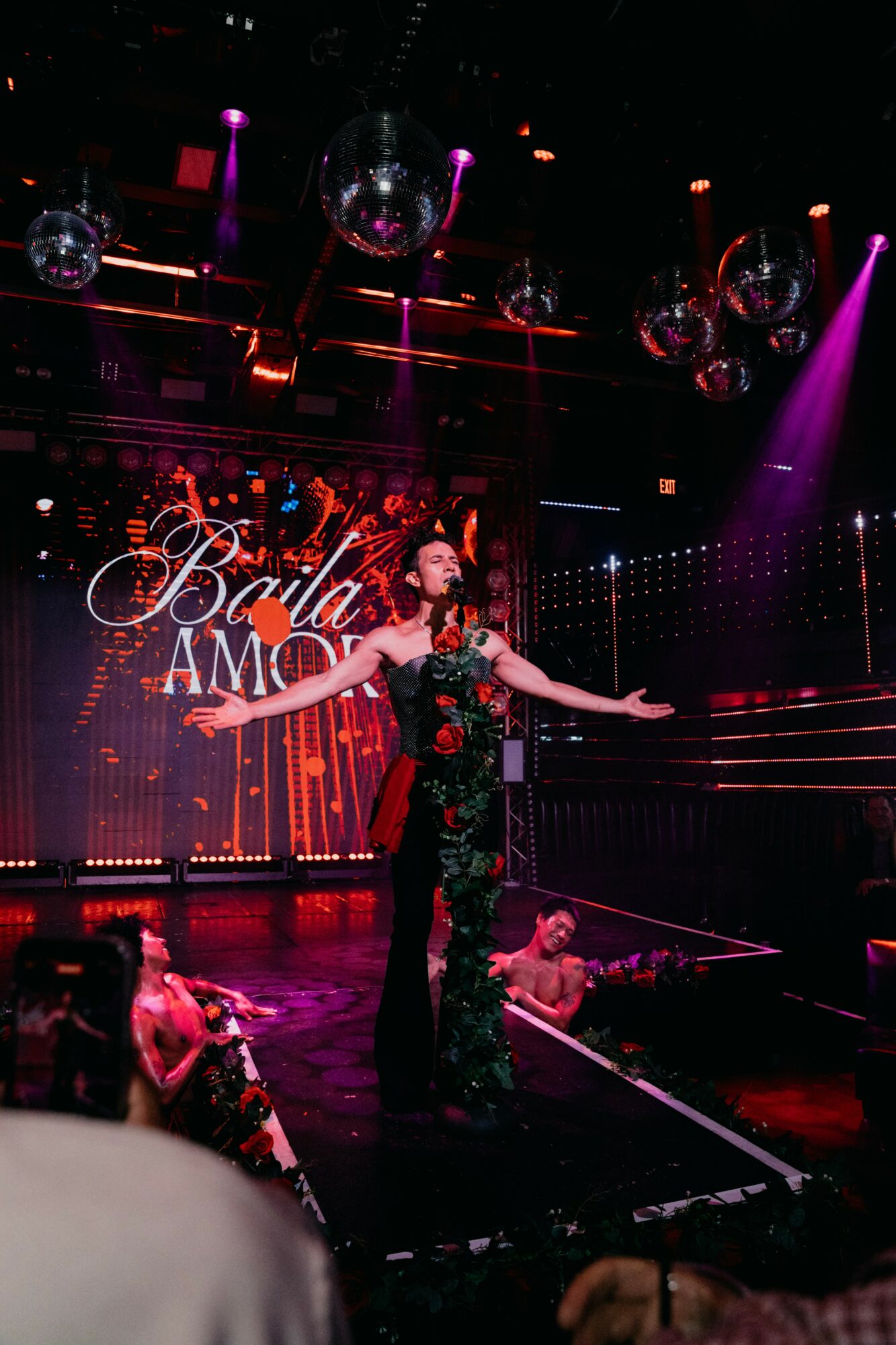 Performer on stage with arms outstretched, colorful lights, and a backdrop reading 'Baile AMO', in a lively nightclub setting.
