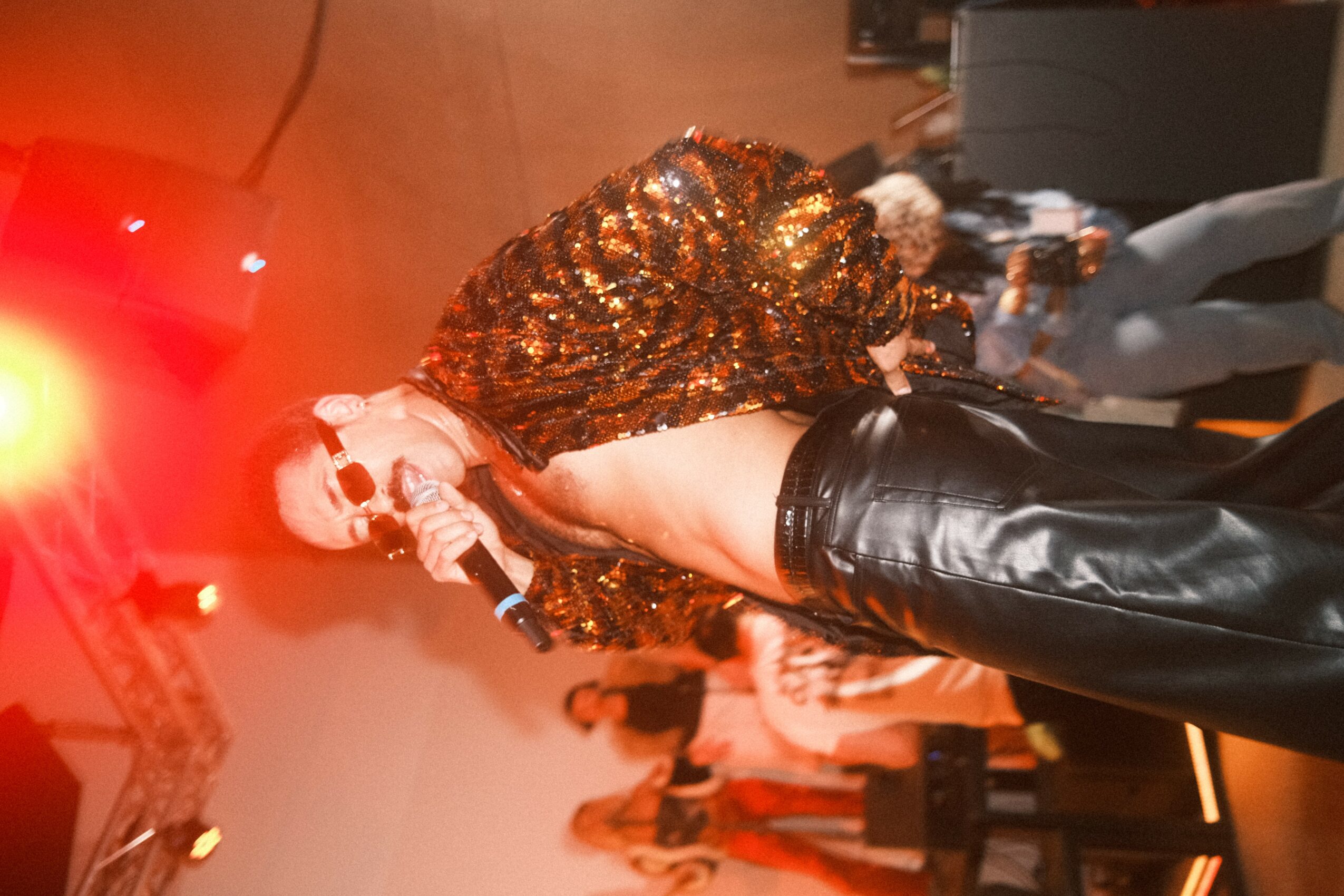 Performer on stage wearing a shiny, sequined jacket and leather pants, with a microphone in hand, under red stage lighting.