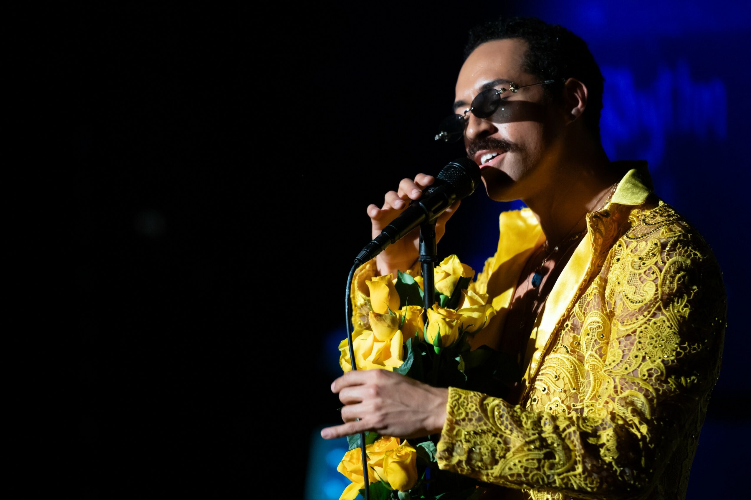 Person singing into a microphone, wearing sunglasses and a yellow patterned jacket, on stage with dark background.