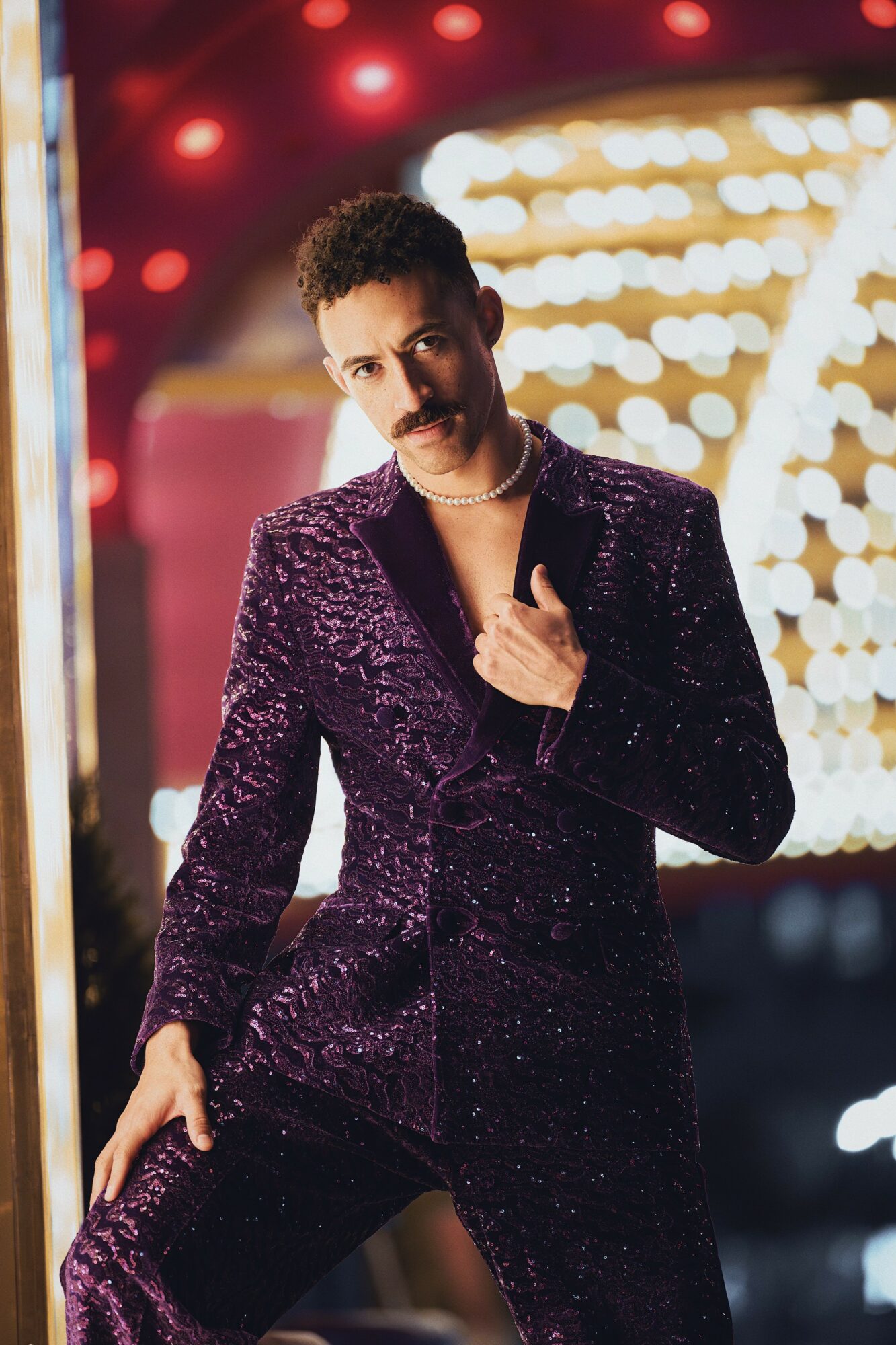 Person in a shiny purple suit standing in front of bright lights, looking at the camera, hand near chest.
