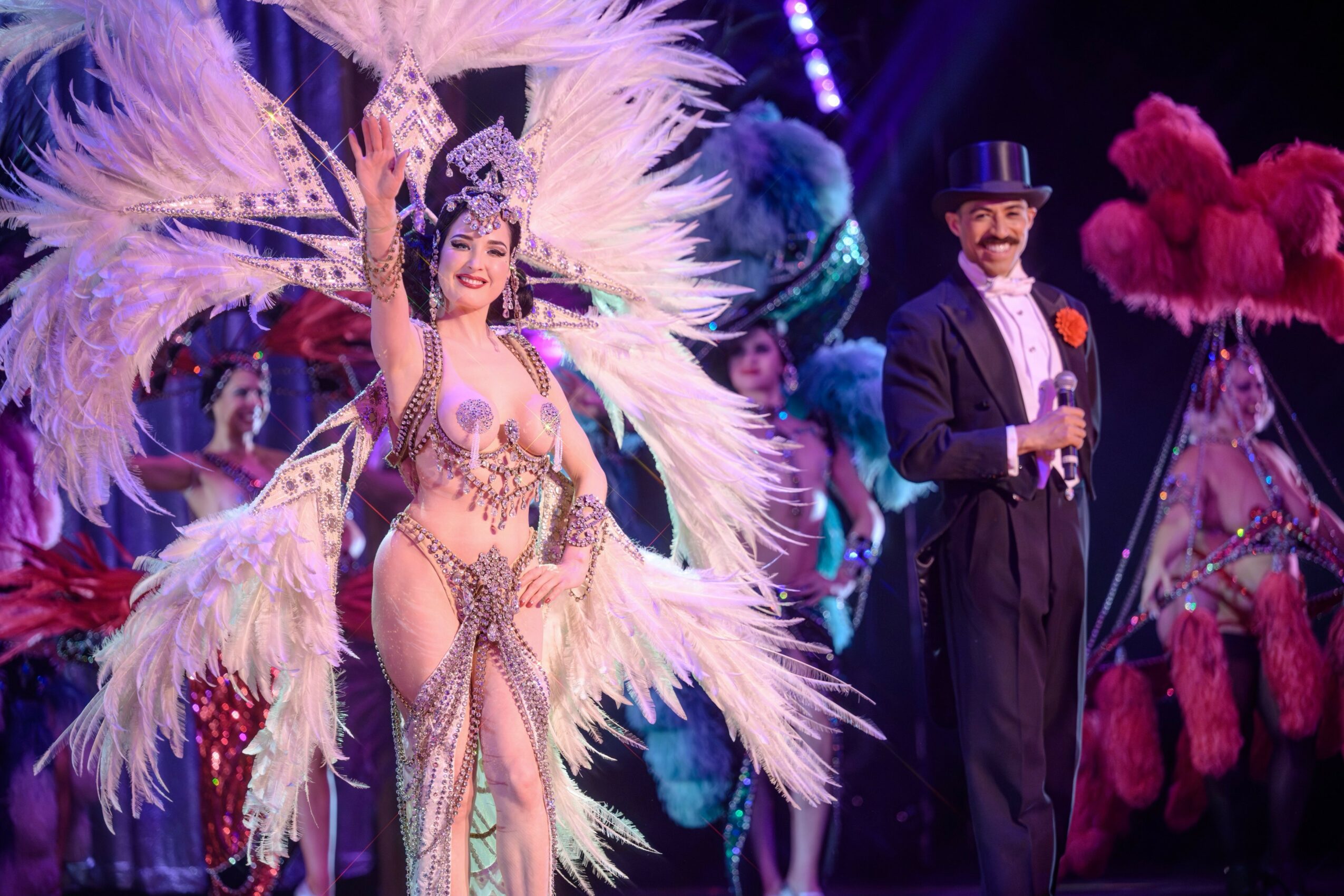 Performer in elaborate feather costume and headdress, and a man in tuxedo with top hat, on stage with colorful feathers.