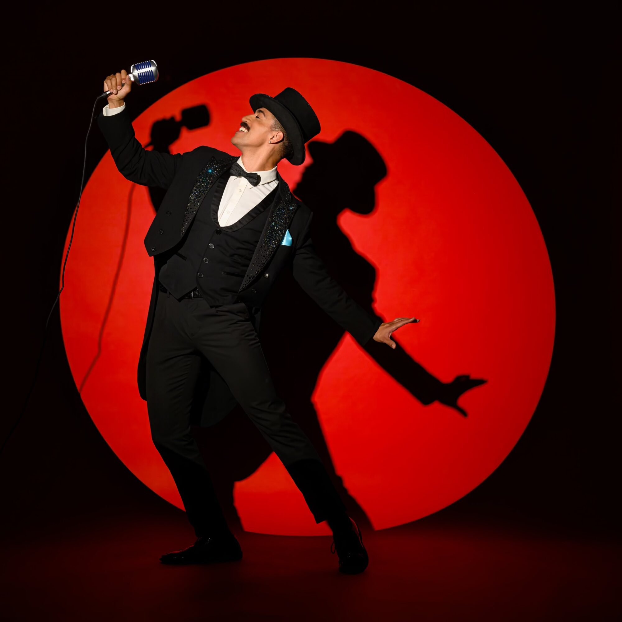 Person in formal attire with a hat, holding a microphone, standing in front of a red circle, performing on stage.