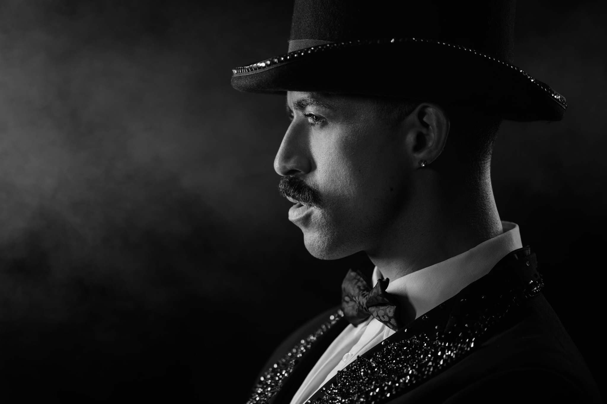 Side profile of a man wearing a hat, tuxedo, and bow tie, in black and white.
