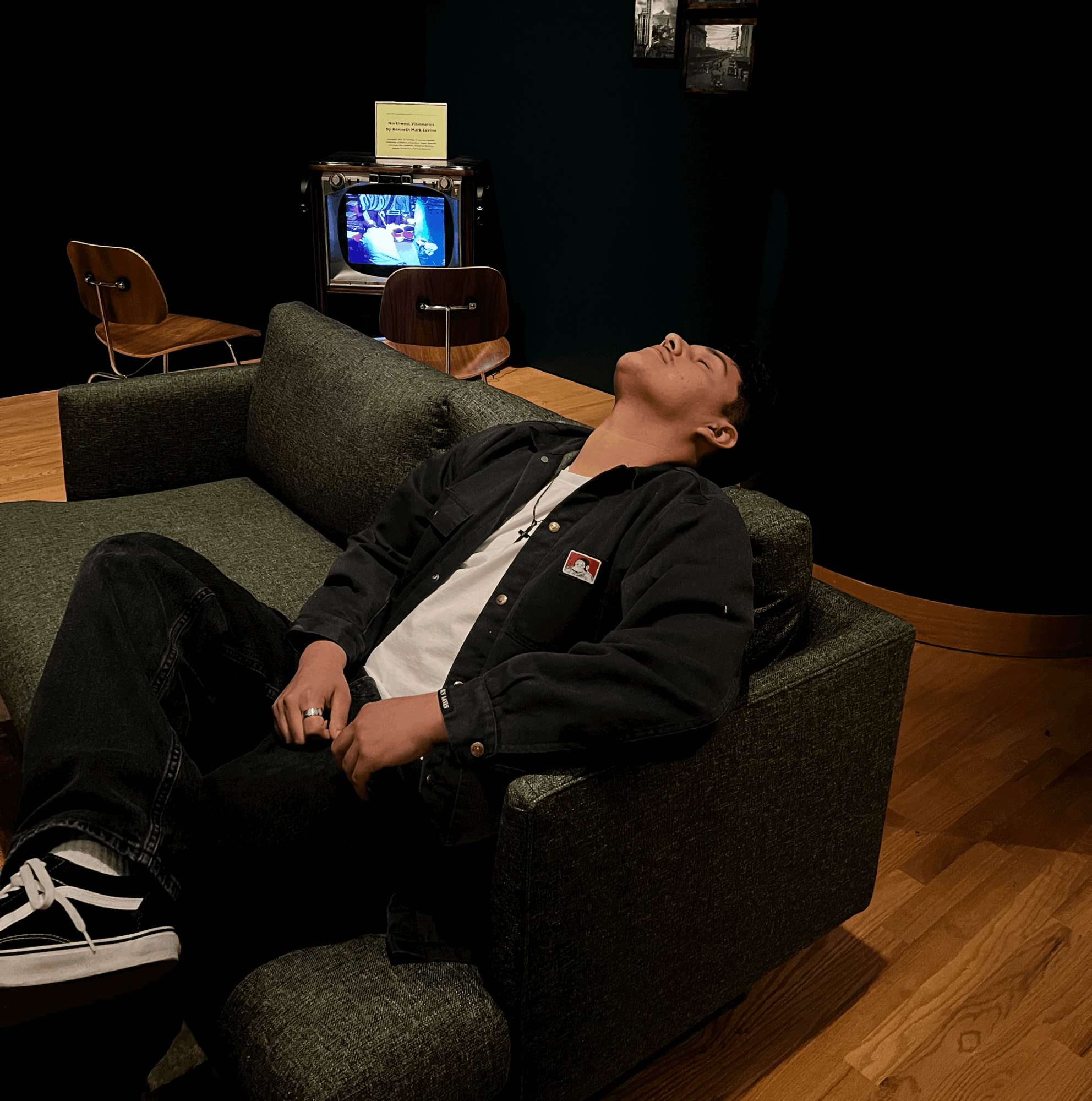 Person reclining on a green sofa with head tilted back, eyes closed, in a room with a TV and chairs behind.