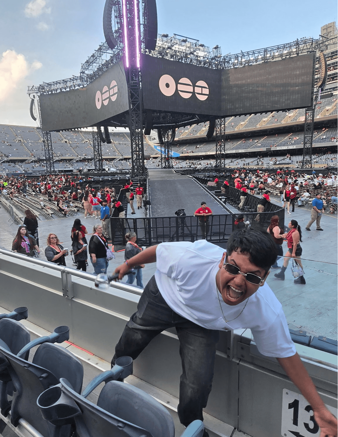 Person leaning over stadium railing, smiling, with a large screen and crowd in the background.
