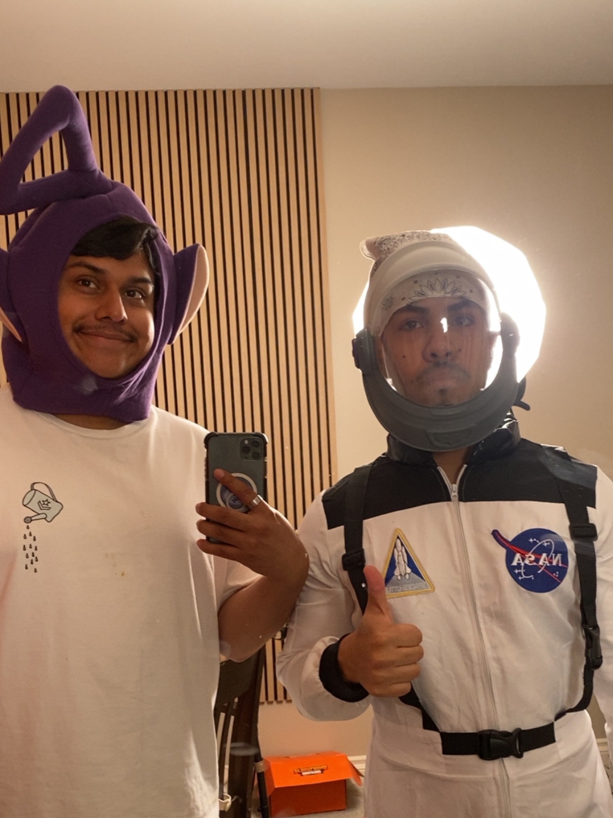 Two people in astronaut costumes taking a selfie in a room with wooden paneling, one giving a thumbs-up, smiling.