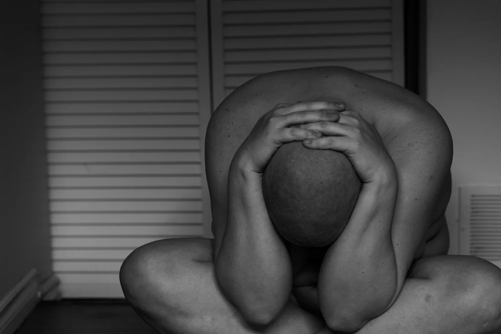 Person sitting with head in hands, arms covering head, in a room with closed blinds, black and white photo.