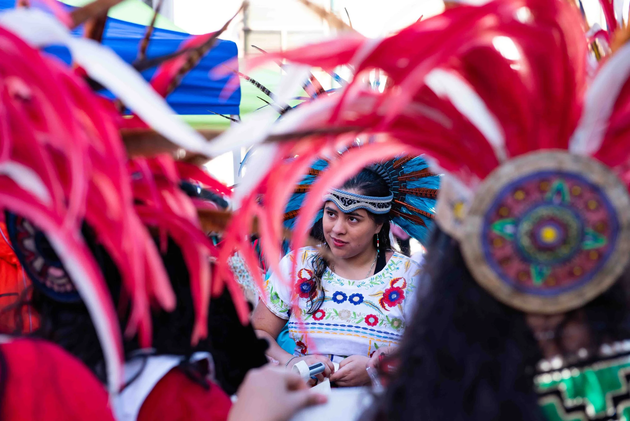 Young woman in traditional colorful attire and headdress, surrounded by people in elaborate costumes with feathers, at a cultural event.