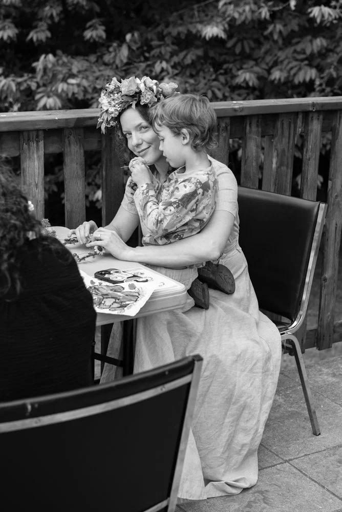 Woman and child sitting at a table outdoors, smiling, with a floral headpiece, surrounded by trees.
