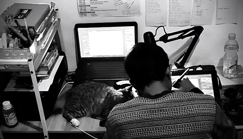 Person working at desk with laptop, books, and a cat lying nearby, in a cluttered workspace, black and white photo.