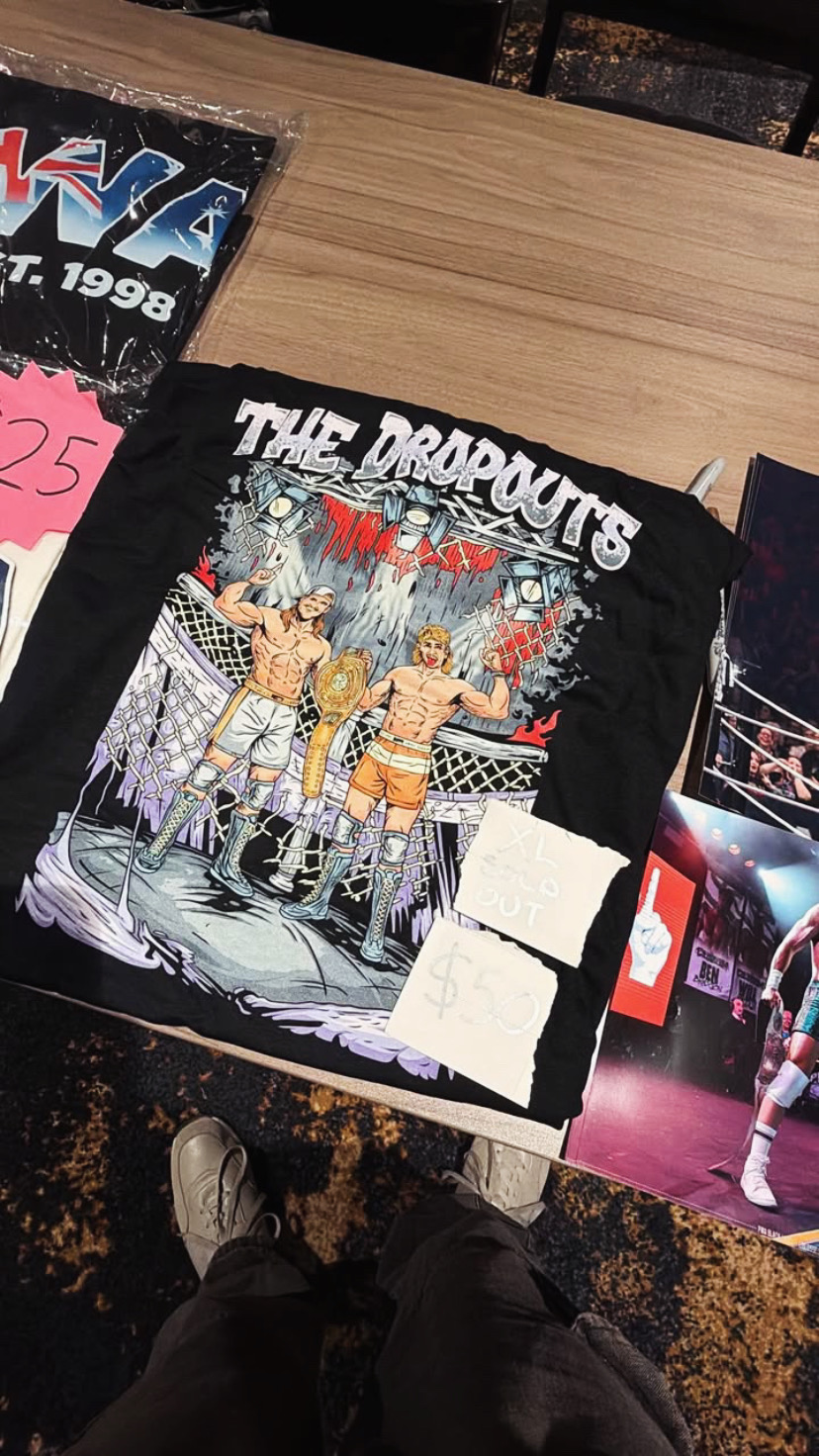 Black T-shirt with a wrestling scene and text, displayed on a table with other items around it.
