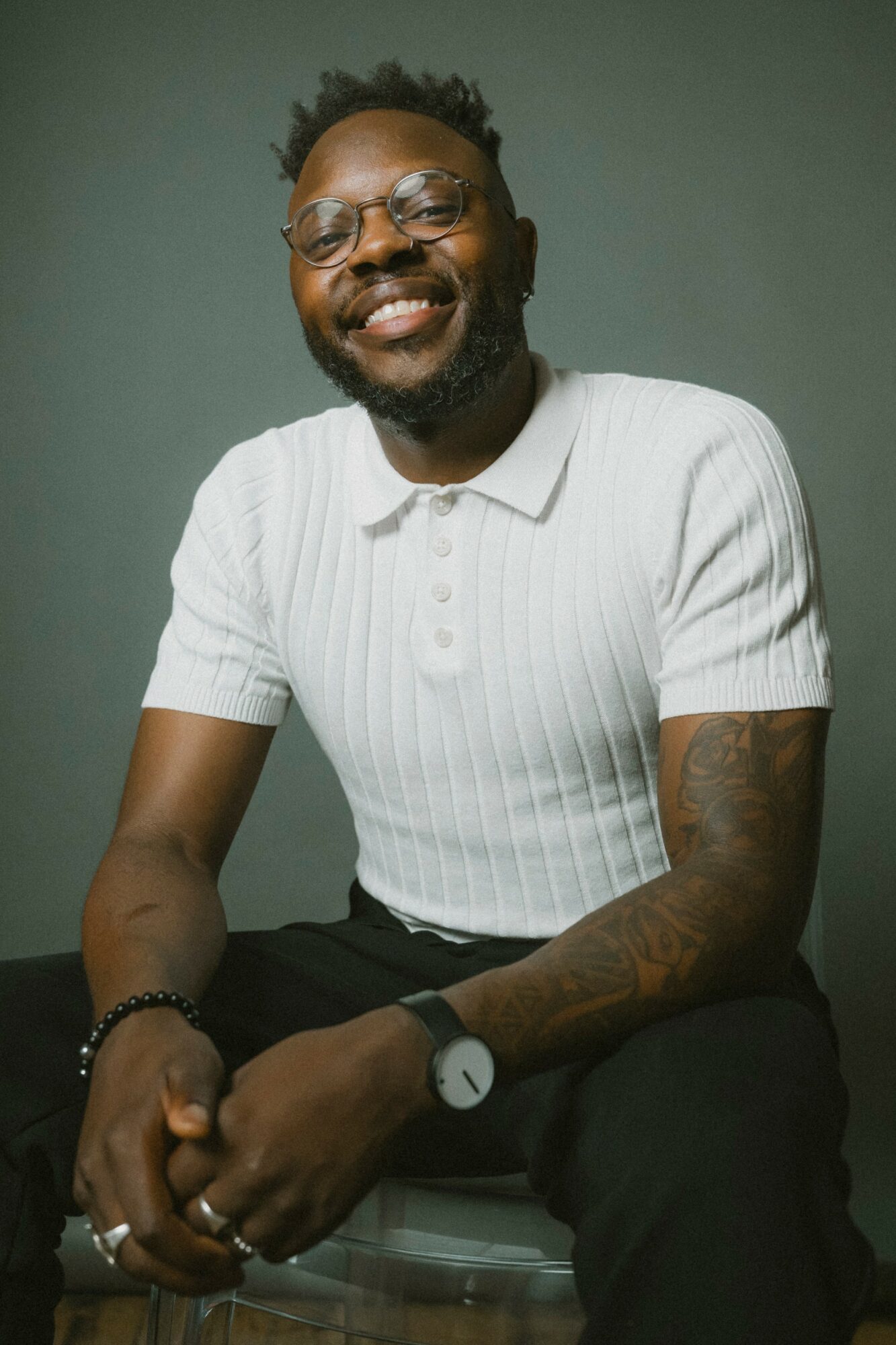 Smiling man with glasses, beard, and tattoos, wearing a white ribbed polo shirt, sitting against a plain background.