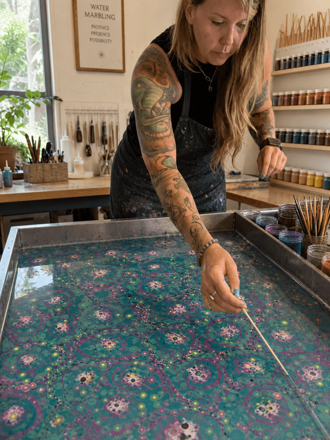 Tattooed woman with long hair leans over a colorful, patterned fabric on a table in an art studio.