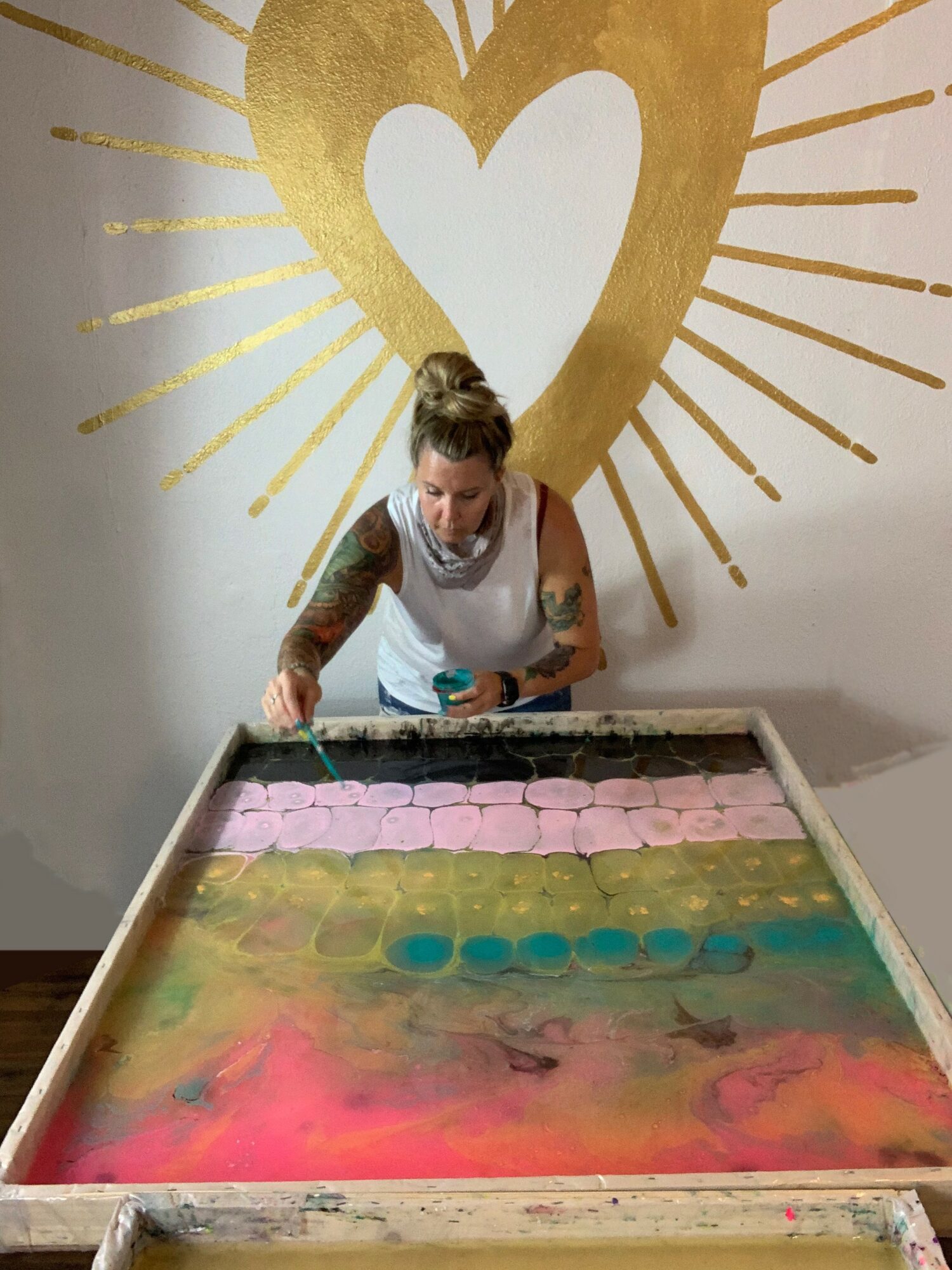 Woman with a bun hairstyle working on colorful soap or wax sheets at a table, with a large heart and radiating lines painted on the wall behind her.