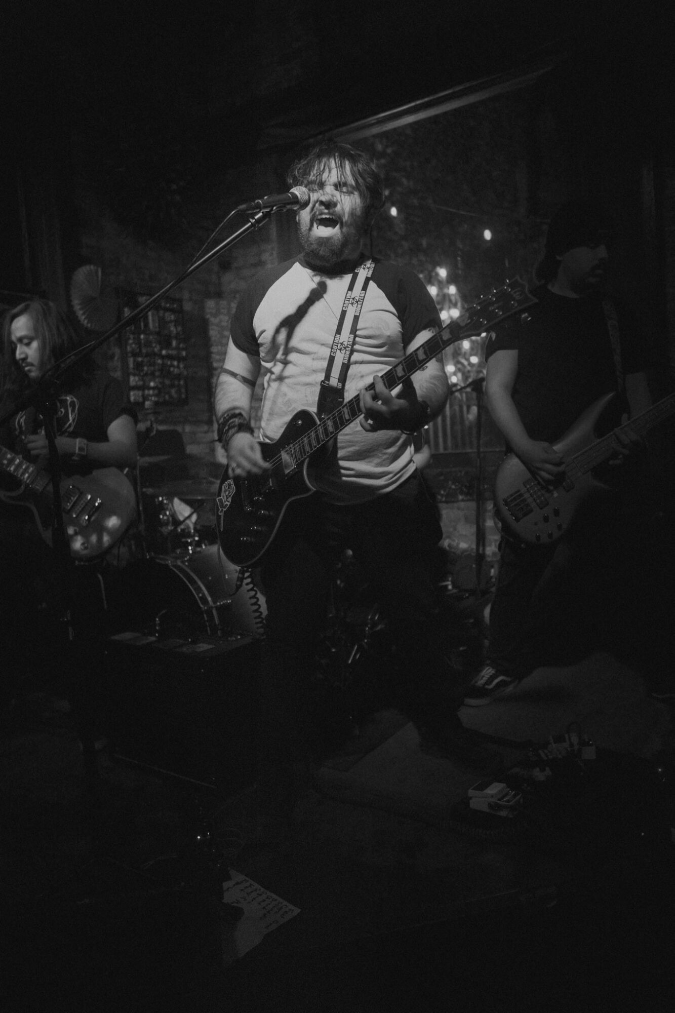 Musician singing and playing guitar on stage with band members, dark background, stage lights, black and white photo.