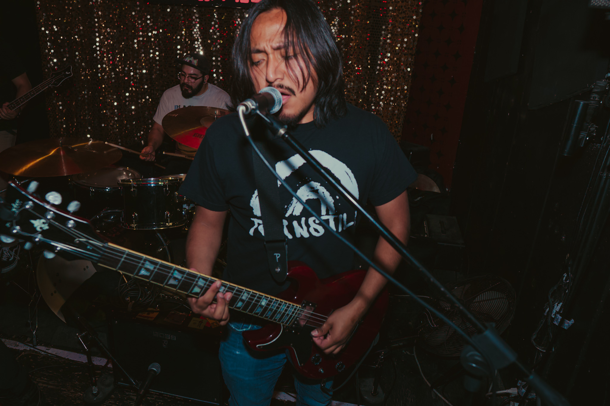 Person singing into microphone while playing guitar, with drummer in background, in a dimly lit setting.