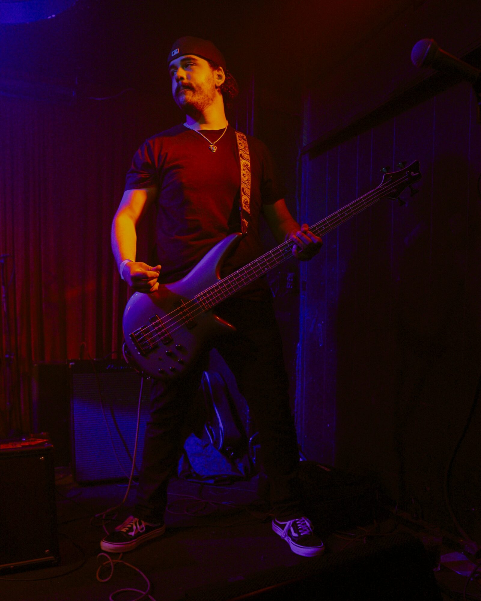 Person playing bass guitar on stage with dark background, wearing a black shirt and cap, illuminated by stage lights.