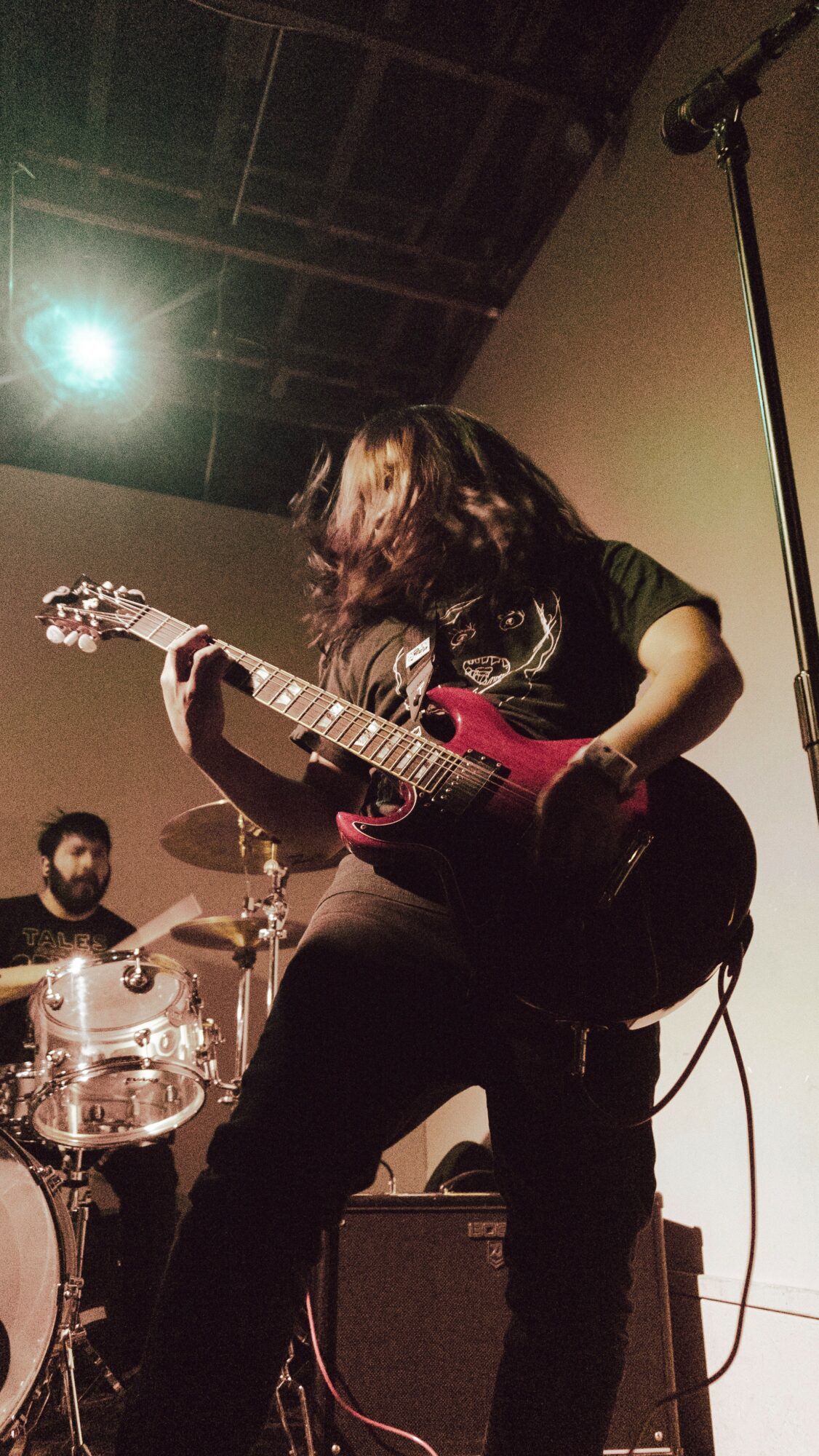 Person playing an electric guitar on stage with a drummer in the background, stage lighting overhead.