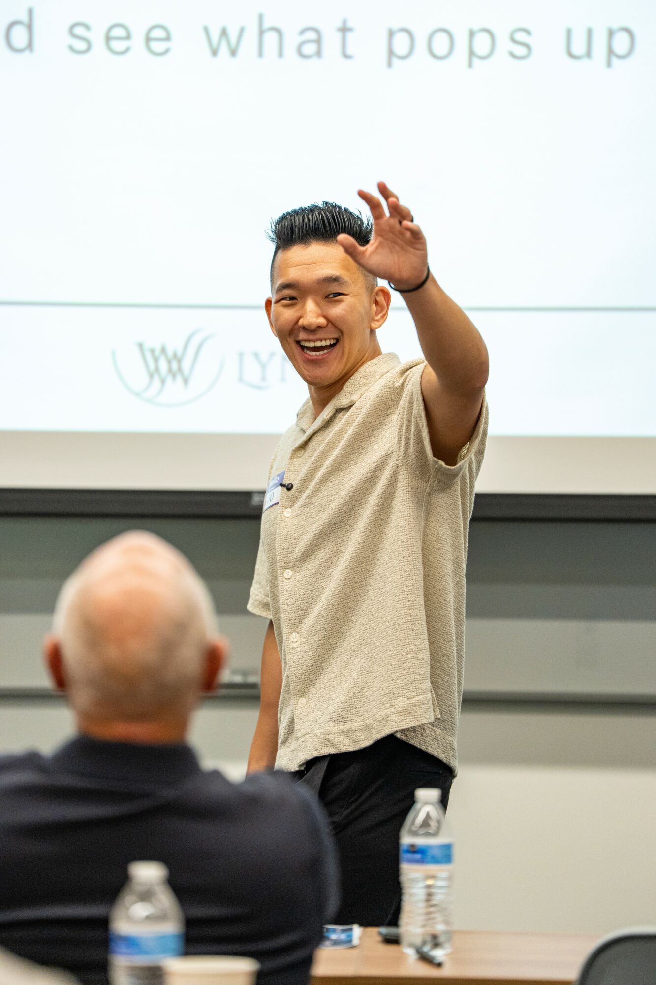 Smiling man waving in front of a presentation screen, with an audience member in the foreground, water bottles on table.