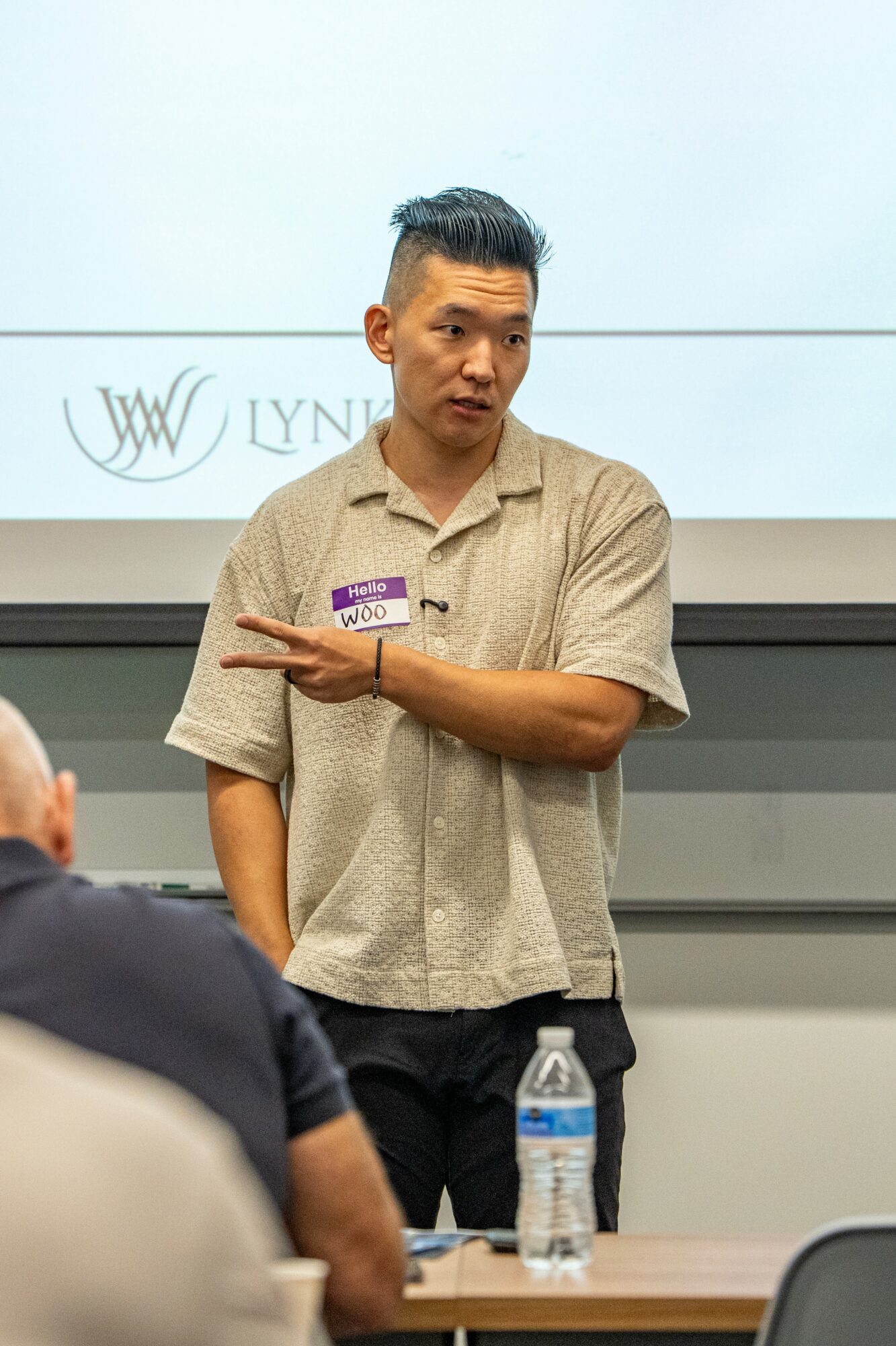 Man standing and speaking in front of a screen, with people seated in a classroom setting, water bottle on table.