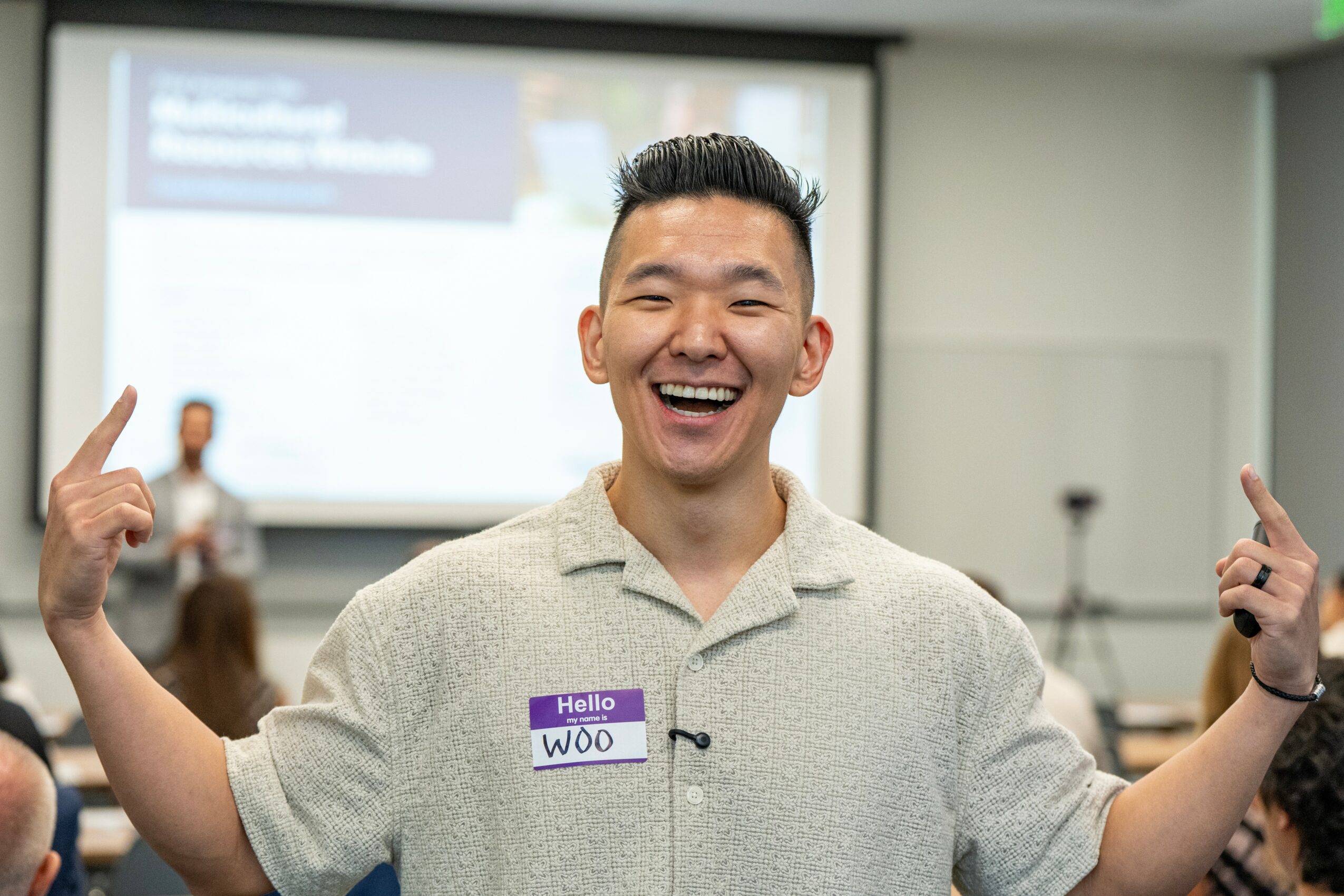 Smiling person with short hair and a name tag, raising both arms in a classroom or conference room.