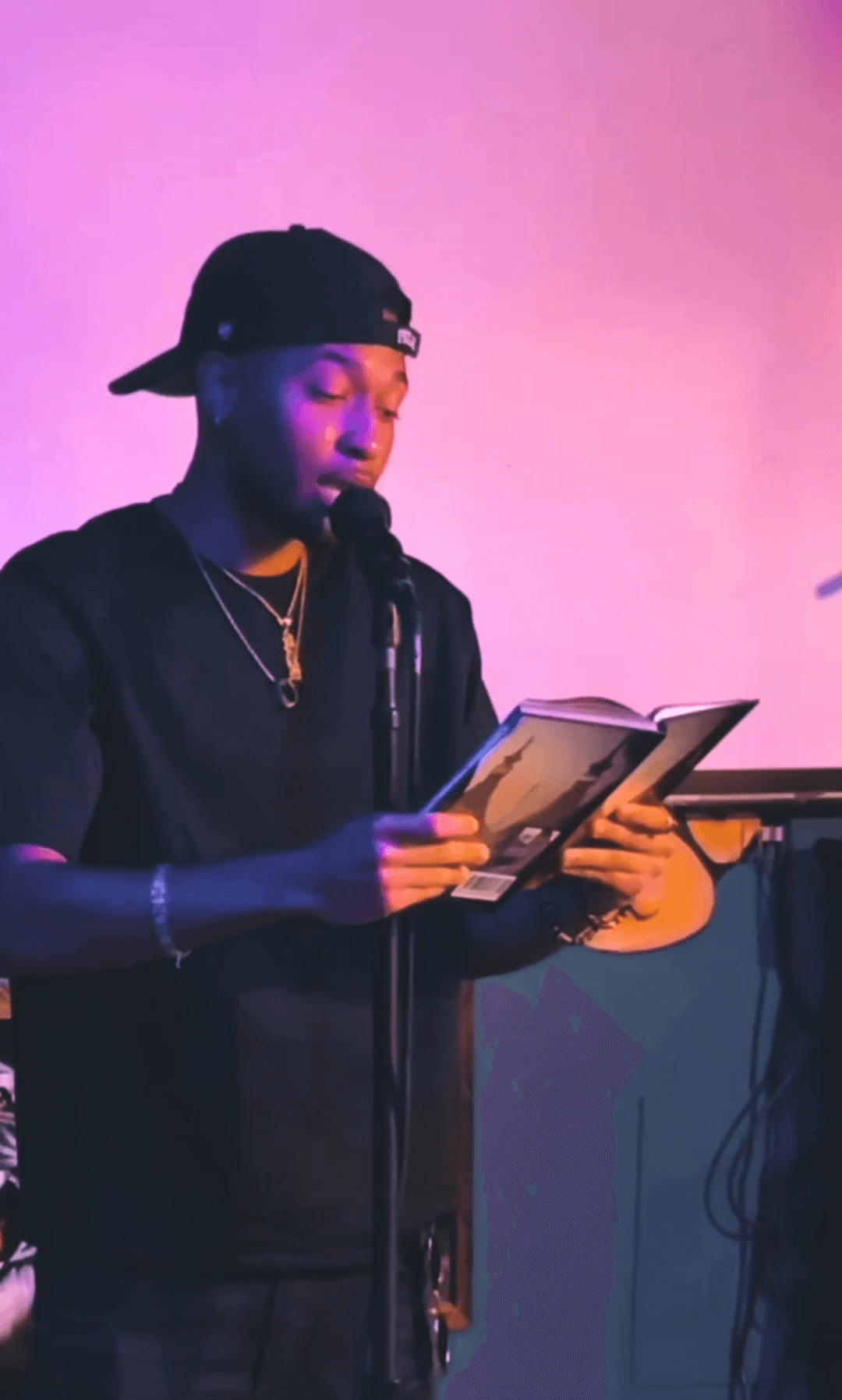 Person wearing a black cap and jacket reading a book at a microphone, pink background, holding a tablet.