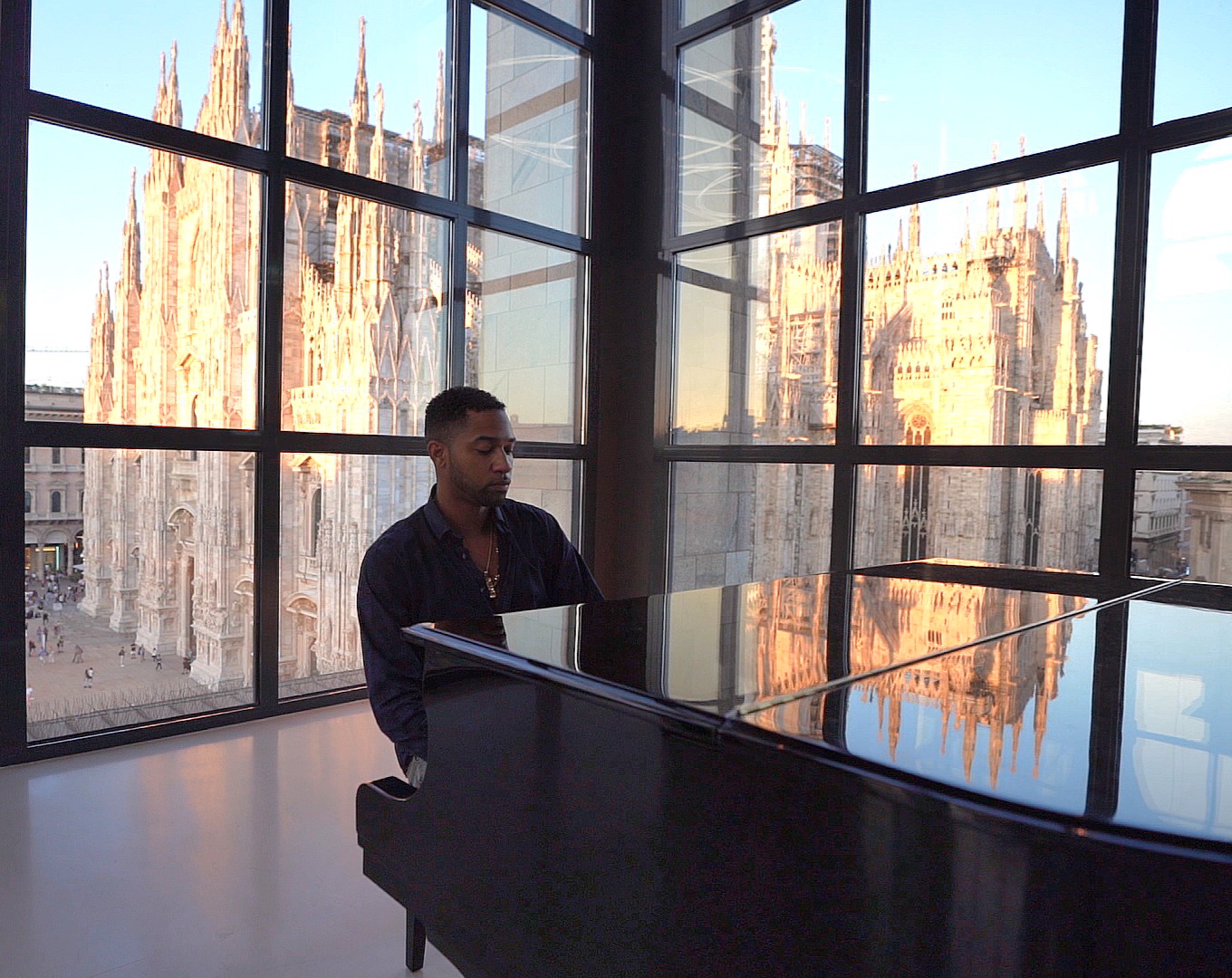 Person playing piano in front of large window with cityscape view, sunlight reflecting on buildings.