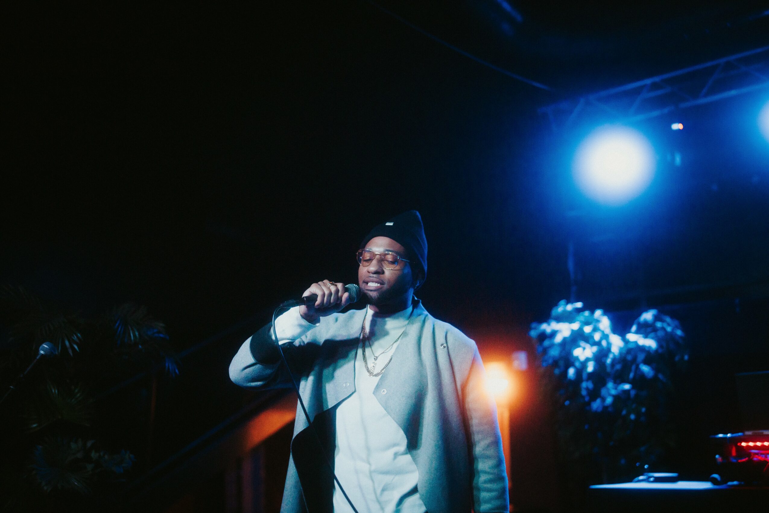 Person singing into a microphone on stage with bright blue and orange lights, wearing glasses and a beanie.