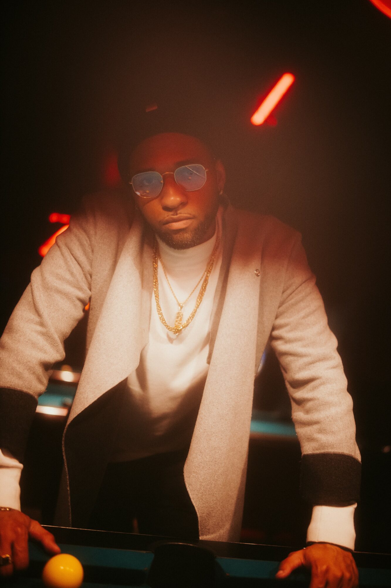 Man wearing sunglasses and a beige coat with gold chains, standing at a pool table in a dimly lit setting.