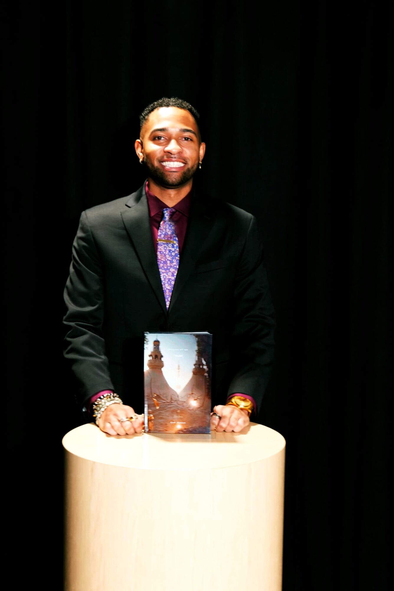 Man in a suit standing behind a table with a book, smiling, against a dark background.