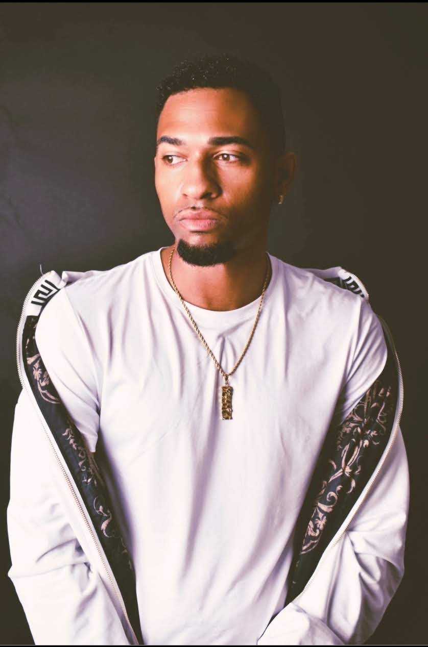 Young man with short hair and a serious expression, wearing a white long-sleeve shirt with black accents, gold necklace, and pendant.