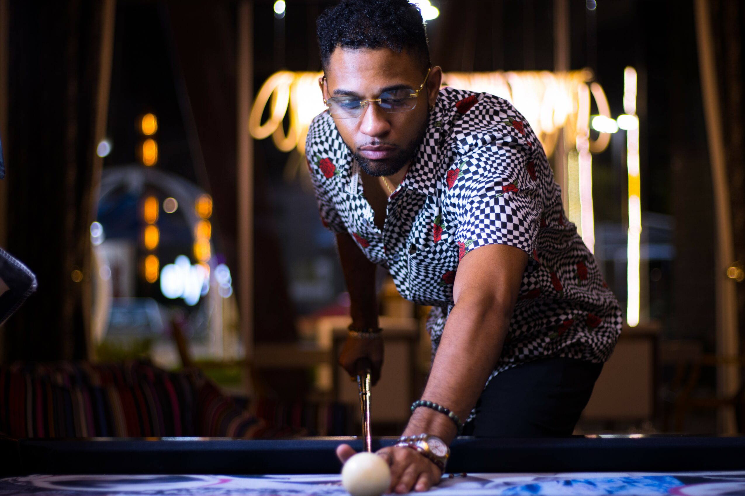 Man playing pool in a dimly lit room with warm lighting and a neon sign in the background.