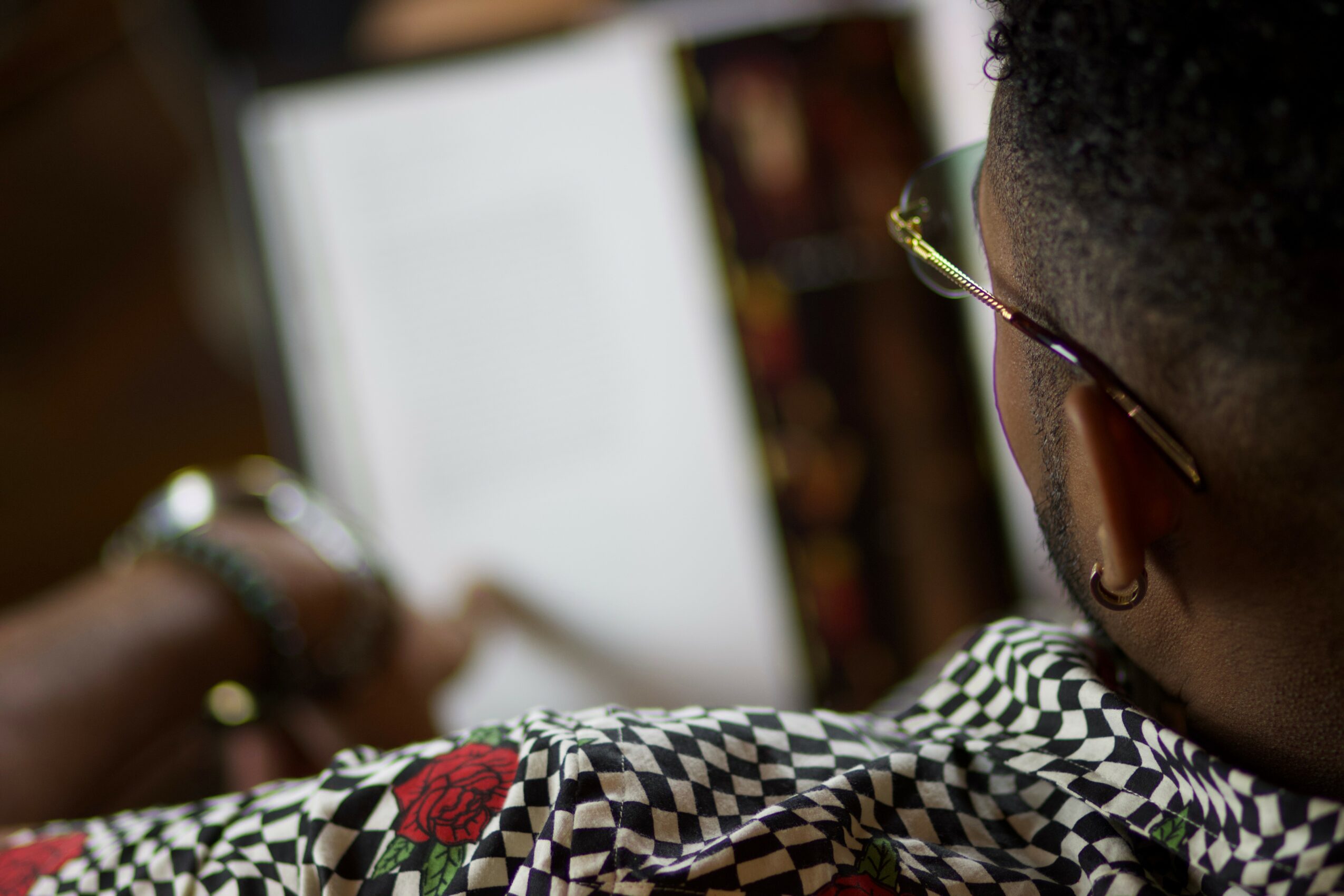 Person with short curly hair and glasses looking at a computer screen, wearing a checkered shirt with a red rose embroidery.
