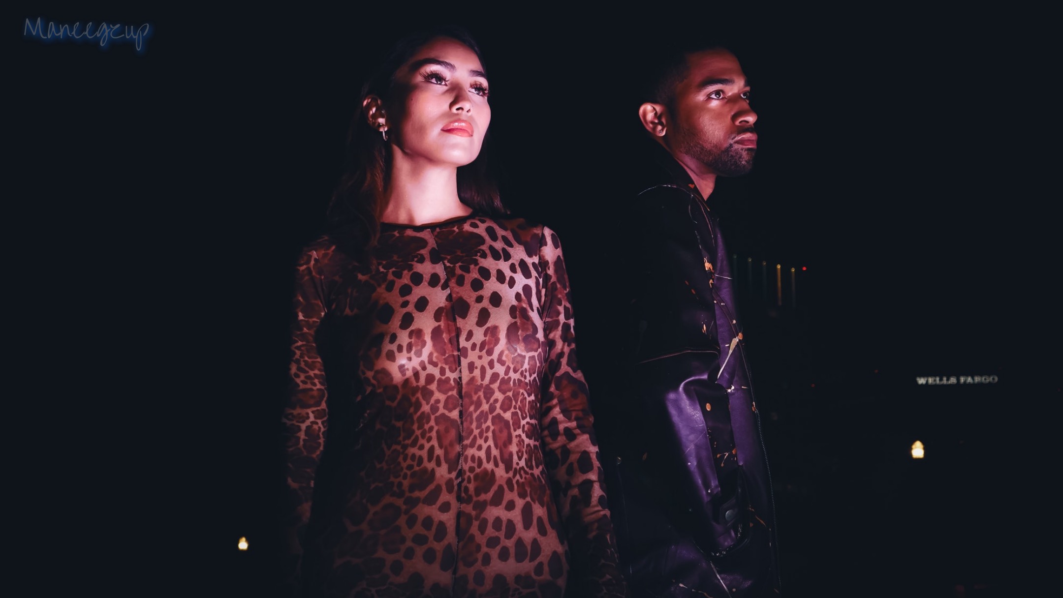 Two people stand side by side in a dark setting, one wearing a leopard print top and the other in dark clothing.