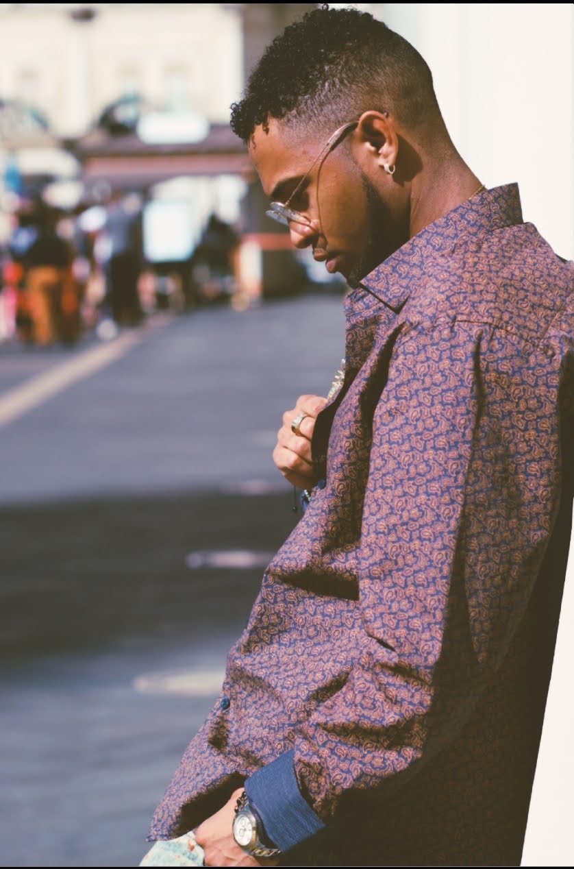 Young man with sunglasses and patterned shirt standing outdoors, looking down, with blurred cars in background.