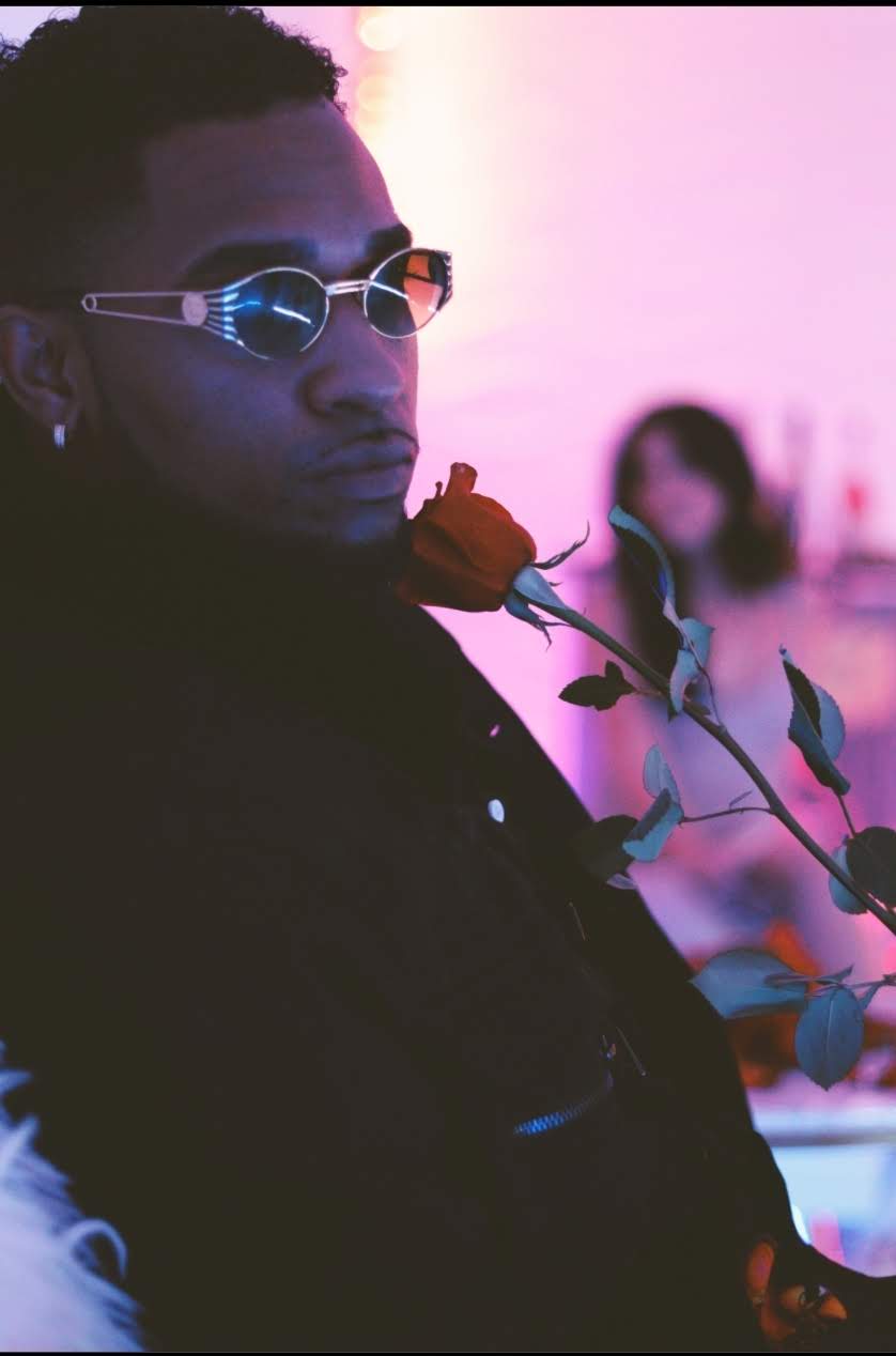 Person wearing sunglasses holding a rose in a dimly lit setting with pink and purple lighting, blurred background.