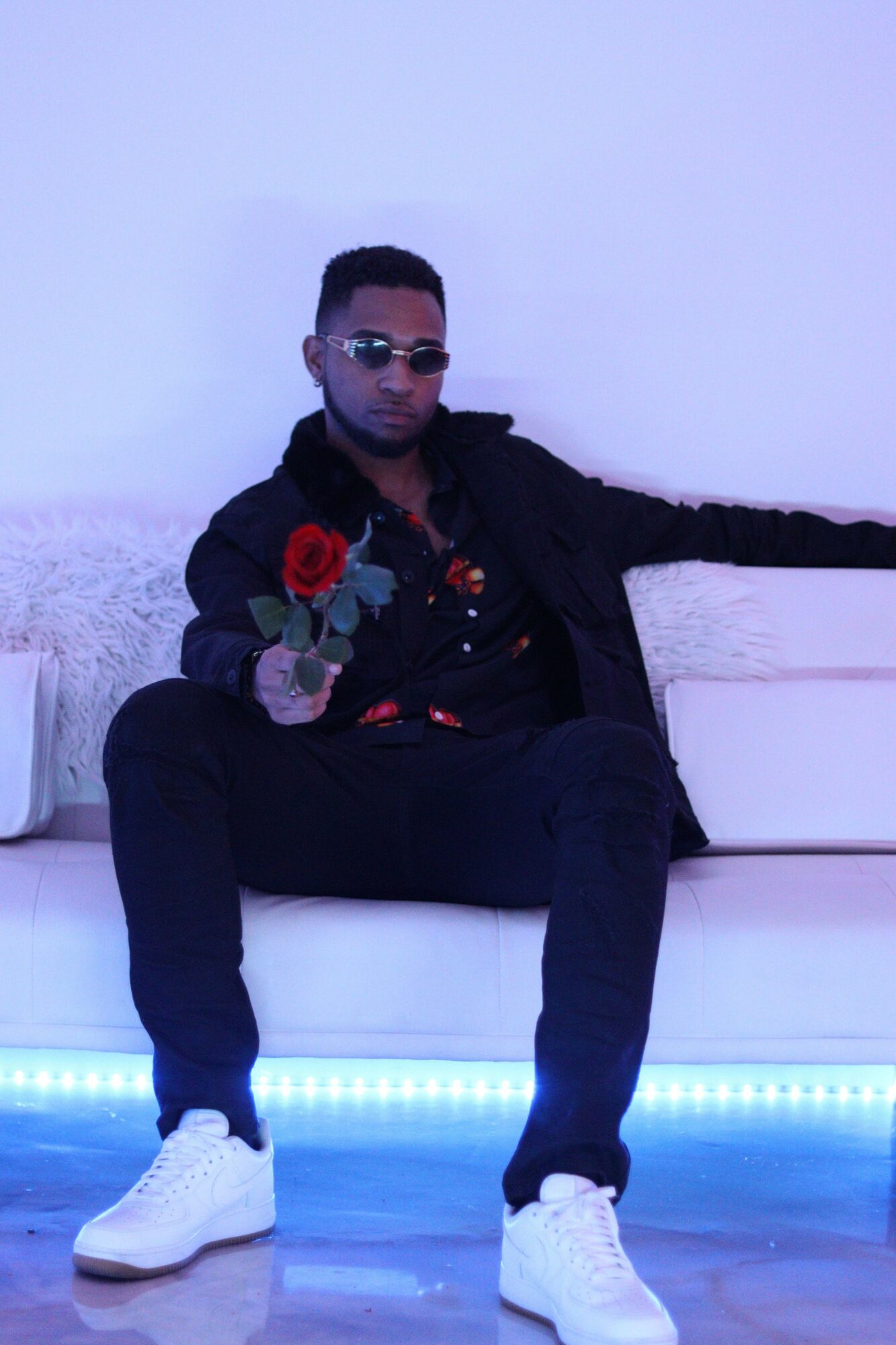 Person sitting on a white couch, wearing sunglasses, black clothing, and white shoes, in a room with blue lighting.