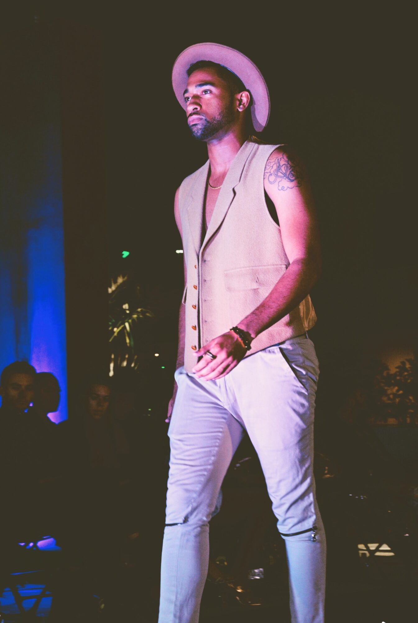 Man with a hat and sleeveless vest walking on stage at night, with audience in background.
