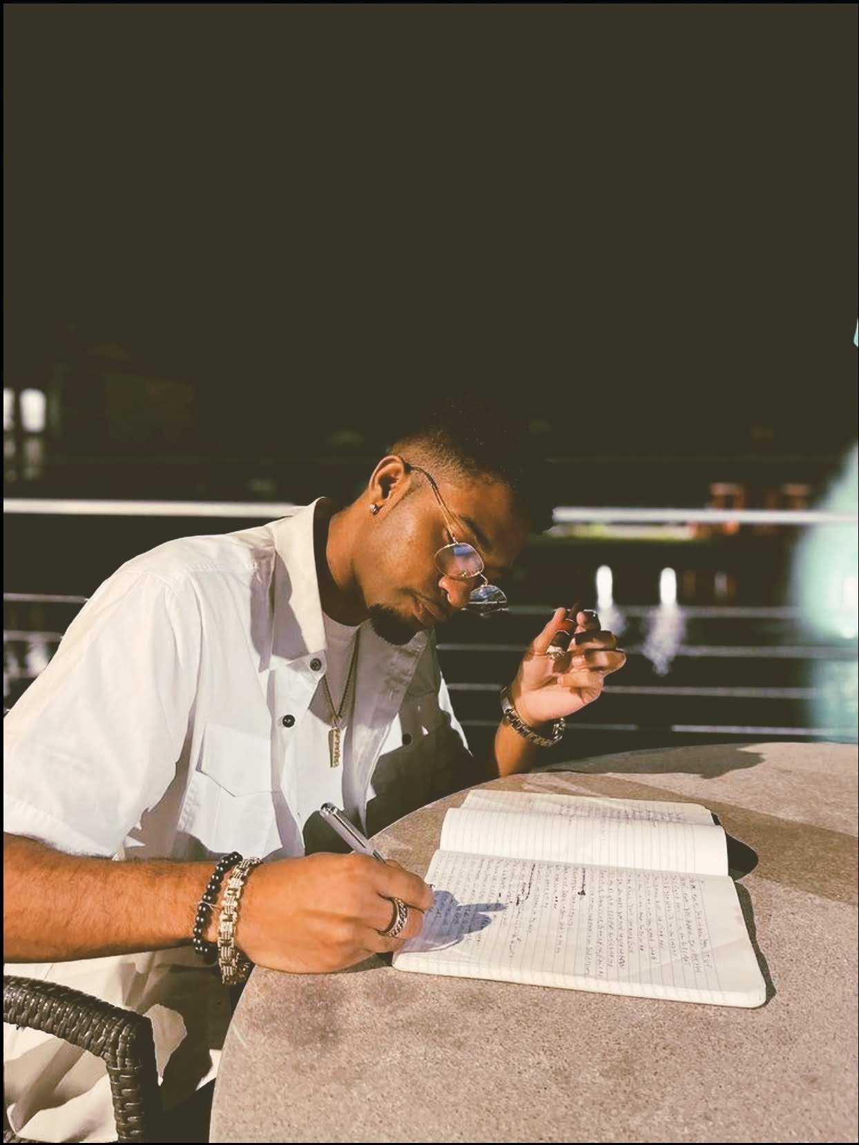 Person reading a book at night, writing with a pen, wearing glasses and a white shirt, sitting at a table.