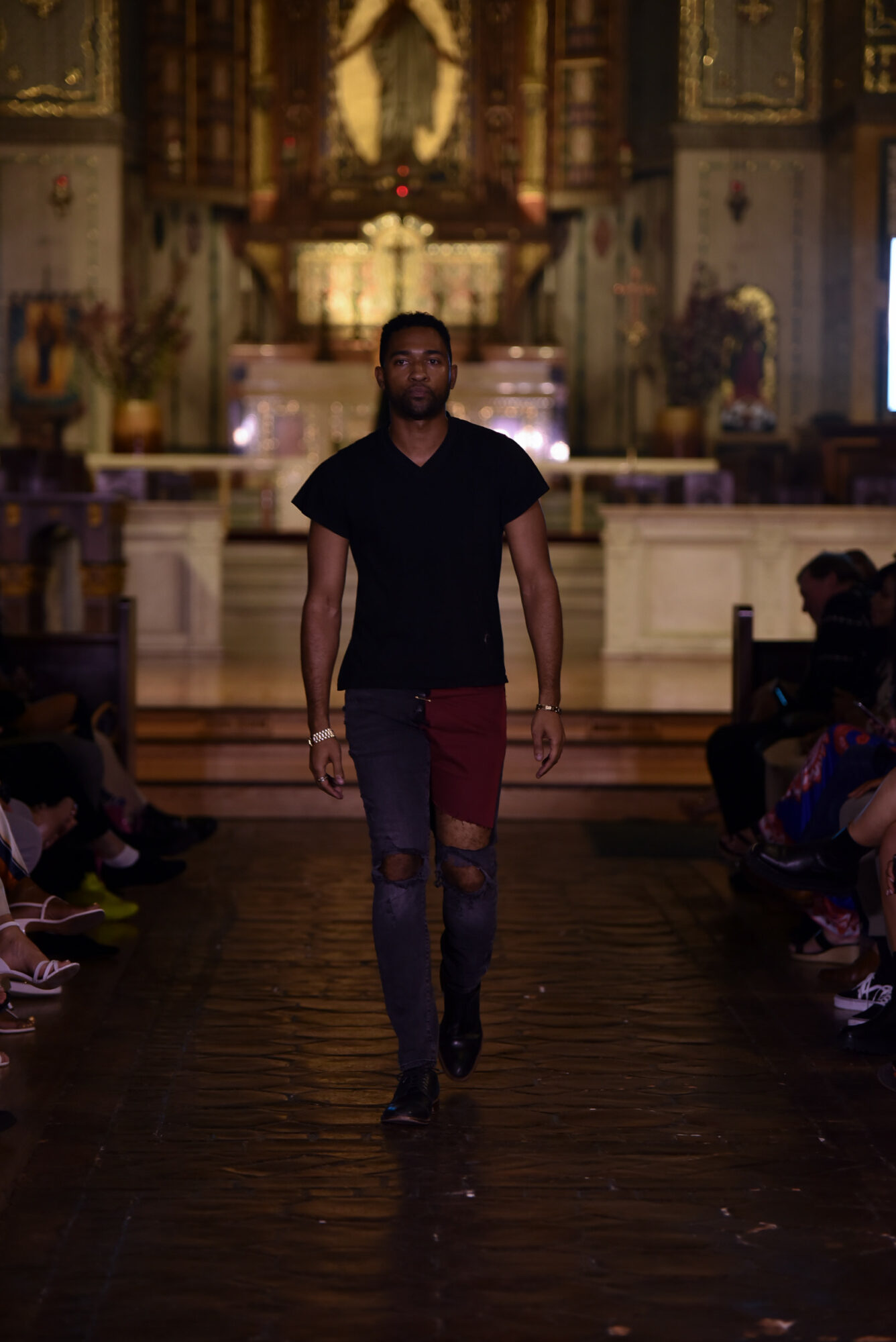 Male model walking down runway in a church-like setting with audience on sides, wearing black shirt and ripped jeans.