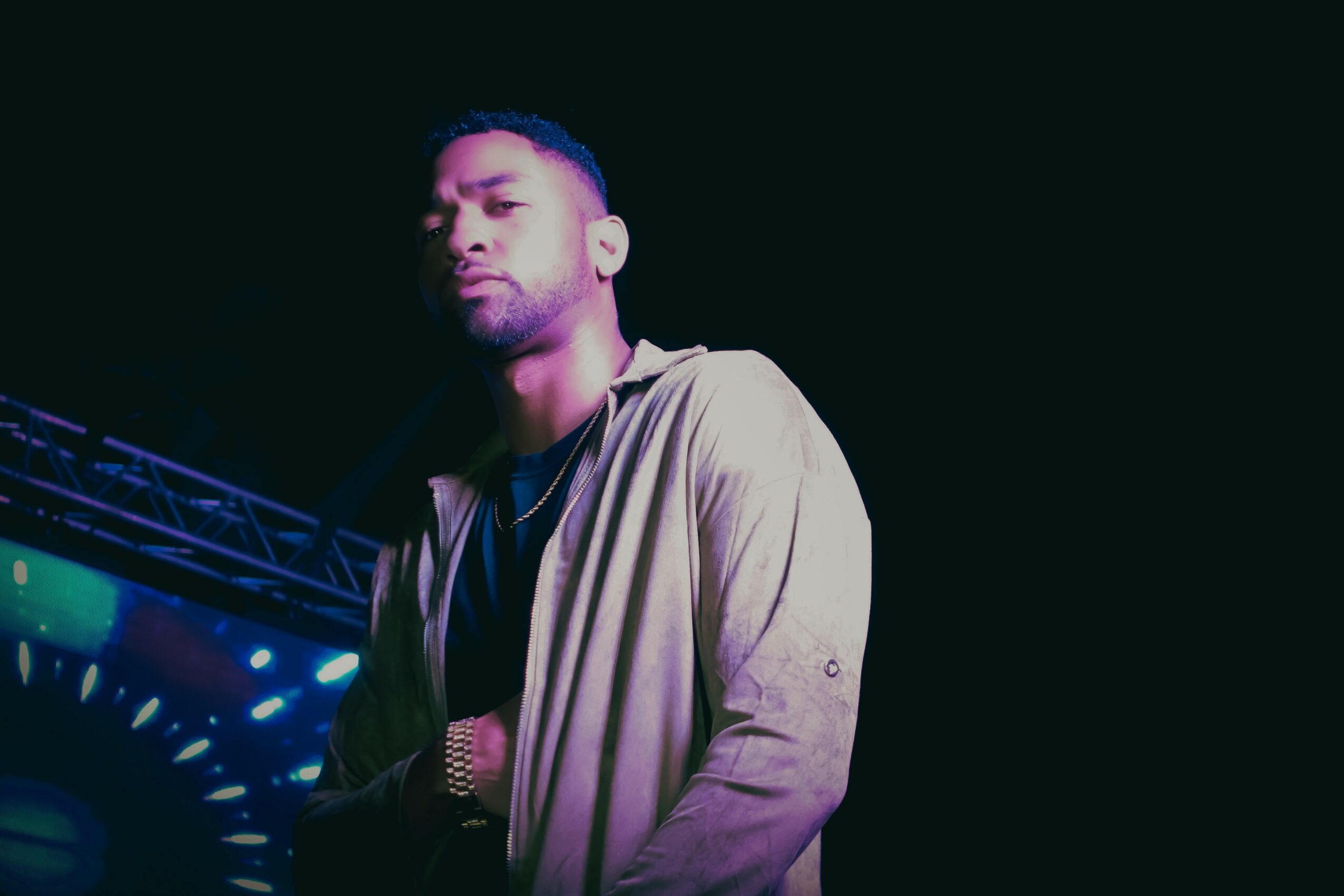 Man with short hair and beard stands with arms crossed, dark background, stage lights visible, serious expression.