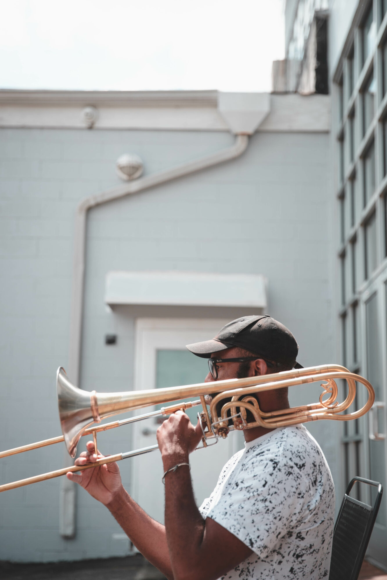Person playing a trombone outdoors, wearing a cap and glasses, with a light-colored building in the background.