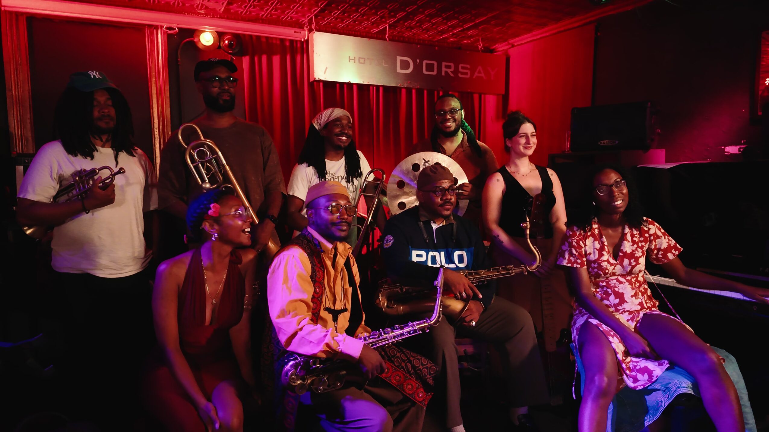 Group of nine people in a dimly lit room with red lighting, some holding musical instruments, posing for a photo.