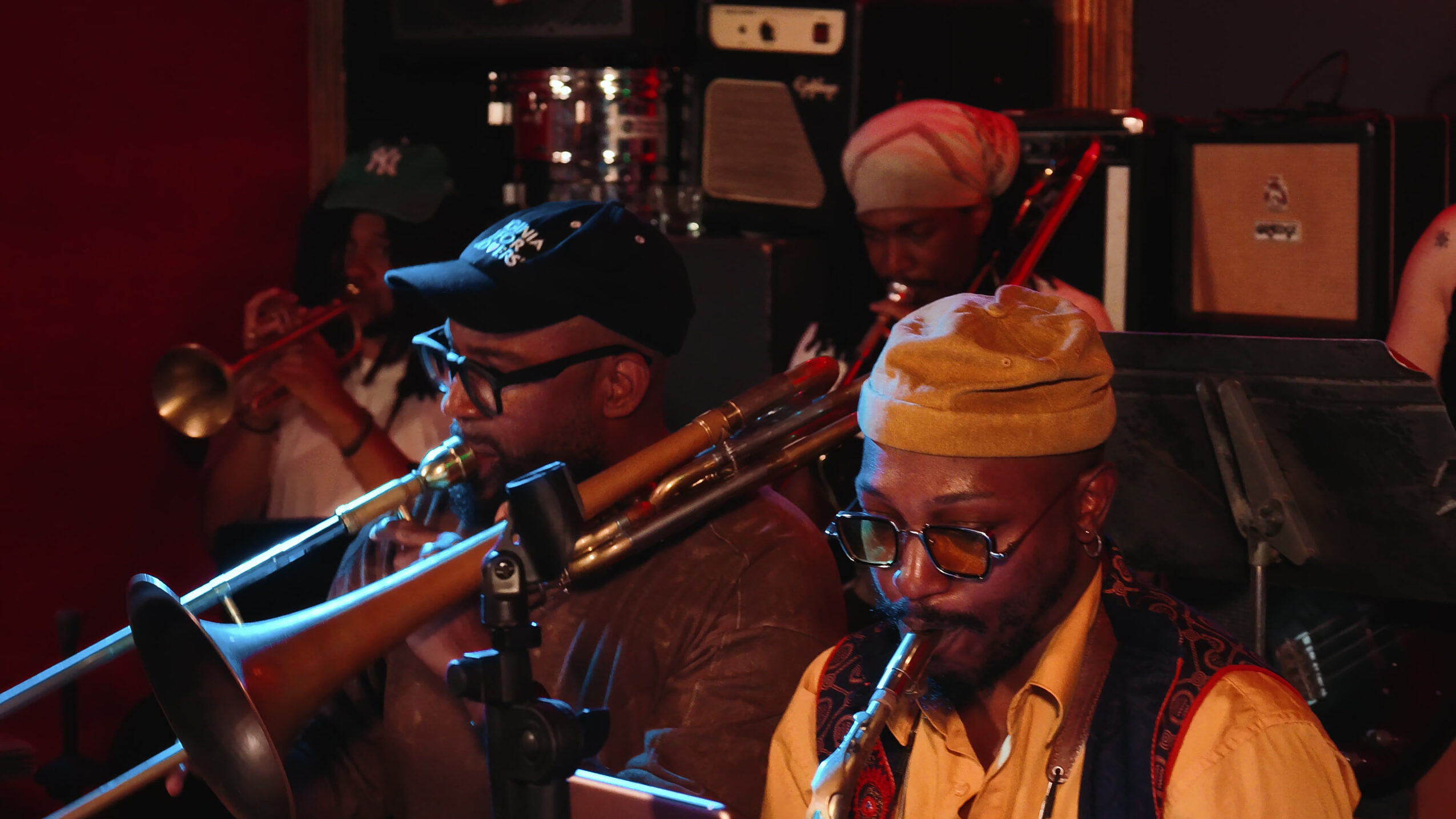 Two musicians playing instruments in a dimly lit room, one with a trumpet and the other with a microphone, wearing glasses and hats.