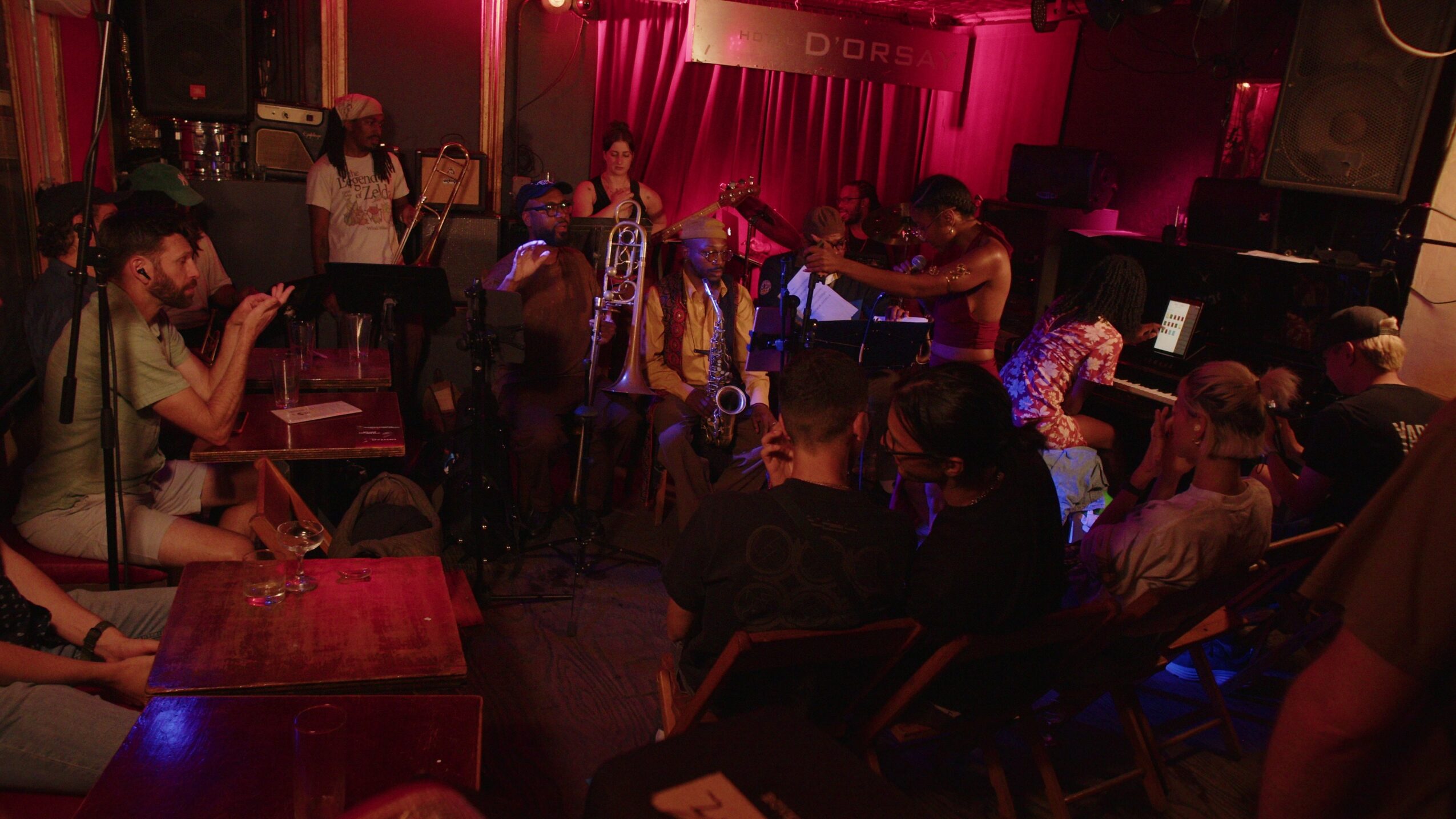 People sitting at tables in a dimly lit venue with a stage and pink lighting, some using phones or talking.