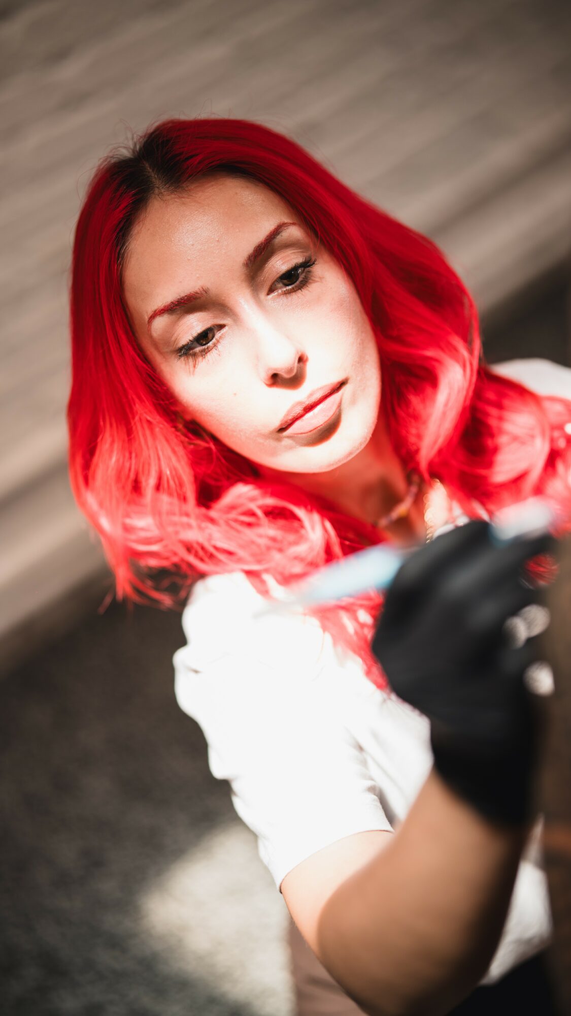 Young woman with red hair looking at a phone, wearing a white top and black glove, blurred background.