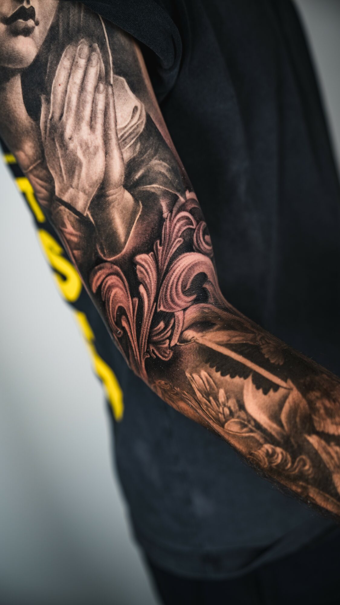 Close-up of a person's arm with detailed black and gray tattoos, including praying hands and floral designs.